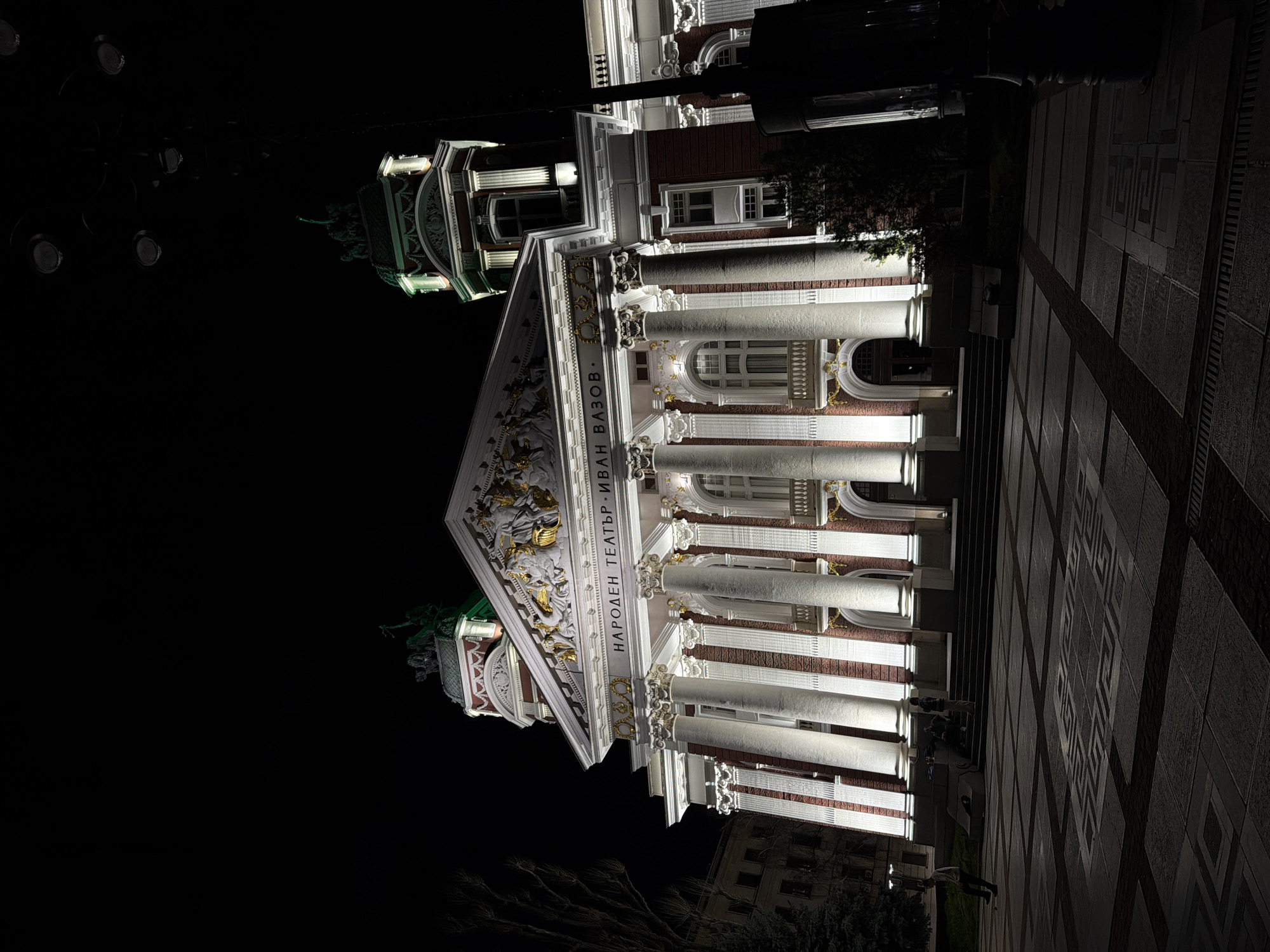 Here is a detailed description of the image:
The image depicts the Ivan Vazov National Theatre in Sofia, Bulgaria, at night. The building is illuminated, showcasing its architectural details such as columns, decorative facade, and statues. The front of the building features prominent white columns with red brick accents. Inscriptions in Bulgarian are visible above the columns. A paved area with geometric patterns extends in front of the building, and a few people are visible walking on the pavement. The background is dark, indicating nighttime. The surrounding environment includes trees and a building with lit windows in the distance. A lamppost with multiple bulbs is present on the left side of the frame.