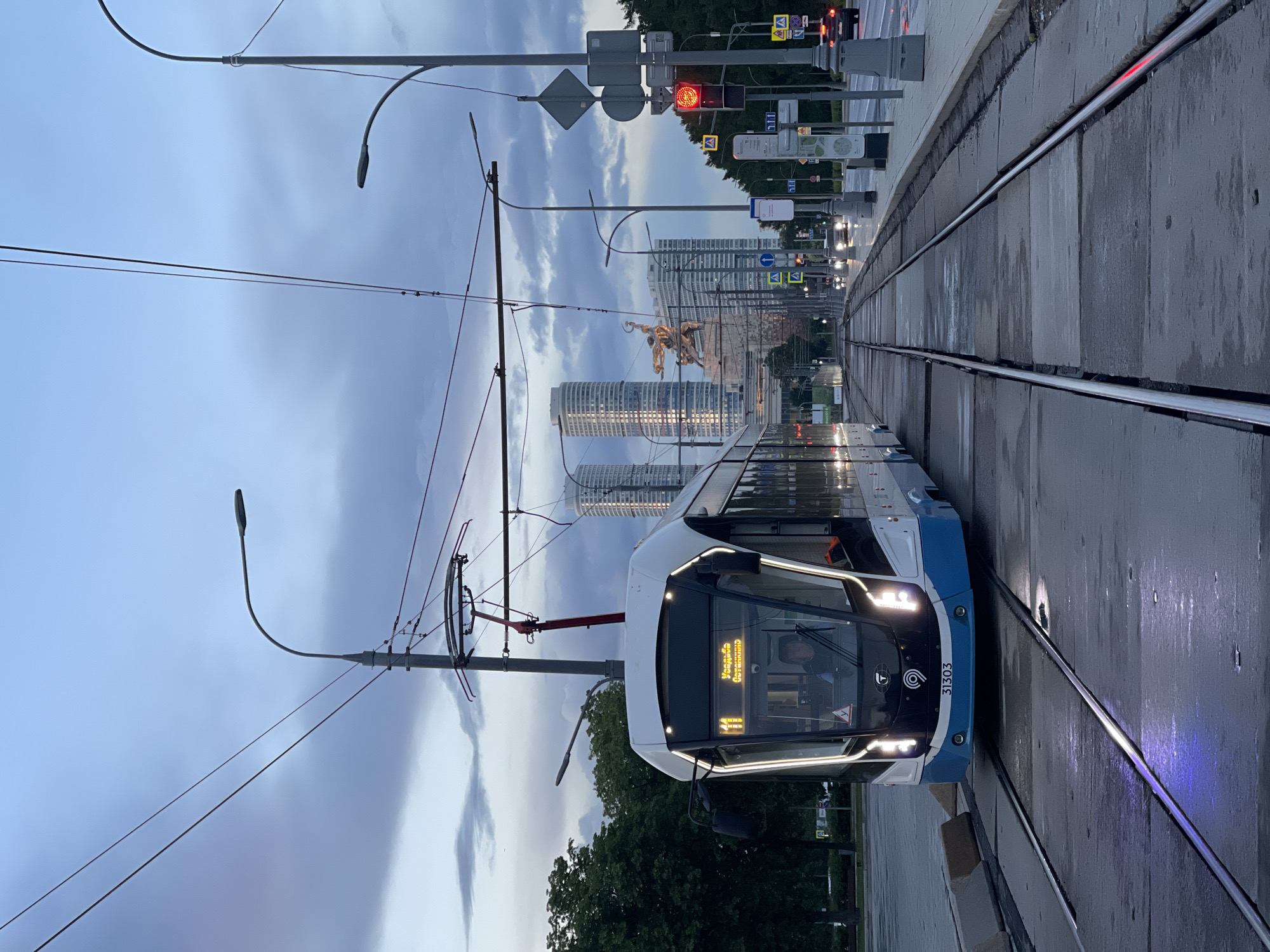 The image captures a modern tram on a track in an urban environment at dusk. The tram, colored white and blue, displays the number "41" and a destination in Cyrillic script ("Улица Седова"). It has its headlights on and appears to be stationary or moving slowly.
The tracks are embedded in concrete, with some water on the surface, suggesting a recent rain. Overhead, there are tram lines suspended by support structures and streetlights. A red traffic light is visible in the background.
In the distance, high-rise buildings and a monument depicting a horse and rider are visible. Based on the architectural style of the buildings and the type of monument, the location is likely to be in Moscow, Russia, possibly near Poklonnaya Gora.
The overall scene evokes a quiet, slightly overcast evening in the city. There's no direct depiction of people interacting within the scene.