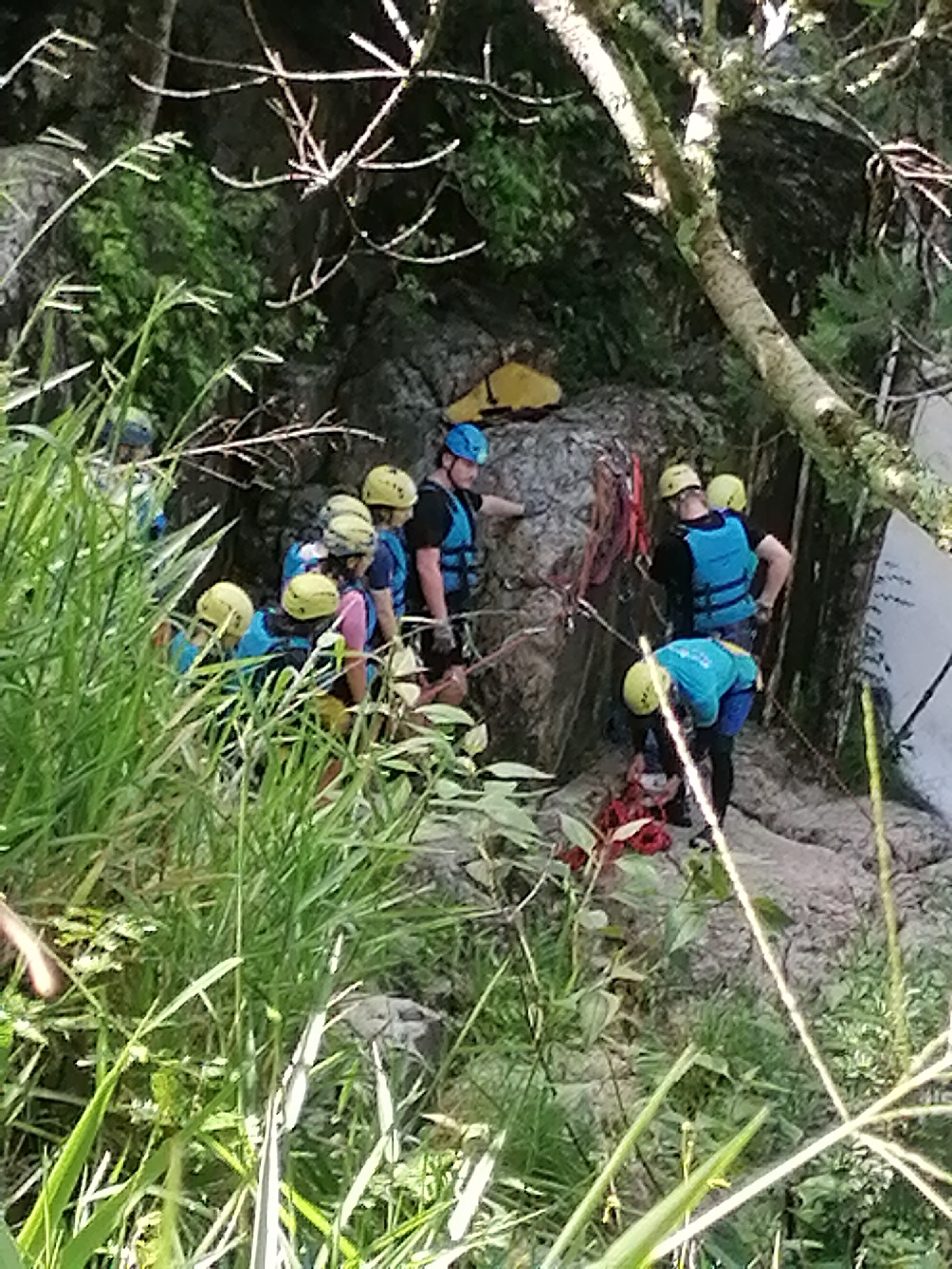 The image depicts a group of individuals engaged in an outdoor activity, possibly a guided excursion or rescue operation, in a rugged, natural environment. Several people are visible, most wearing helmets and life vests in various colors, including blue, yellow, and black. They are positioned on and around a large rock formation, with dense green foliage and trees surrounding them. Ropes and harnesses are evident, suggesting activities such as rappelling or rock climbing. The terrain appears uneven and rocky, with significant vegetation. The lighting suggests it is daytime. The provided context places this scene in Da Lat, Vietnam.