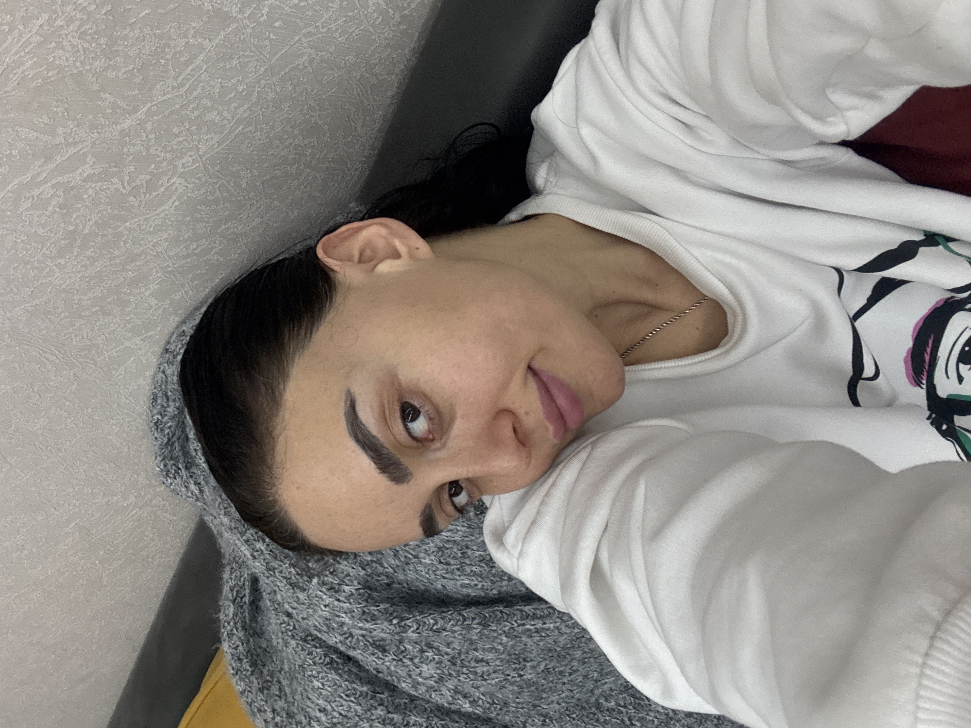 A woman lies on a gray knitted blanket, wearing a white sweatshirt with a cartoon graphic. The background consists of a gray wall. A silver chain necklace is visible around her neck. It is impossible to ascertain the location in Tbilisi, Georgia, from this image.