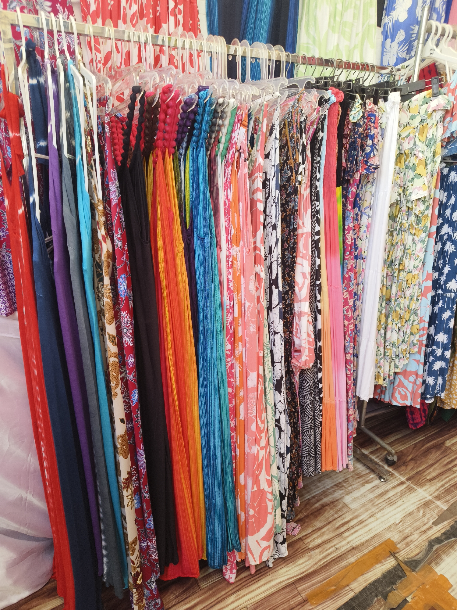 The image depicts a clothing stall in Kuta Selatan, Indonesia, displaying a vibrant array of women's dresses and garments. The items are hung on white plastic hangers from a metal clothing rack, creating a colorful and organized display. The dresses feature a variety of patterns and colors, including floral prints, tie-dye, solid bright hues like red, orange, blue, and purple, and some with intricate traditional Indonesian batik-inspired designs. The background shows more clothing hanging, suggesting a larger retail space. The floor is made of wooden planks with a slightly worn appearance. A metal clothing rack with wheels is visible on the right side of the image, indicating a portable display setup. There are no people visible in the image, and the focus is solely on the merchandise. The overall scene conveys a lively and tropical atmosphere typical of a market or boutique in a tourist destination.