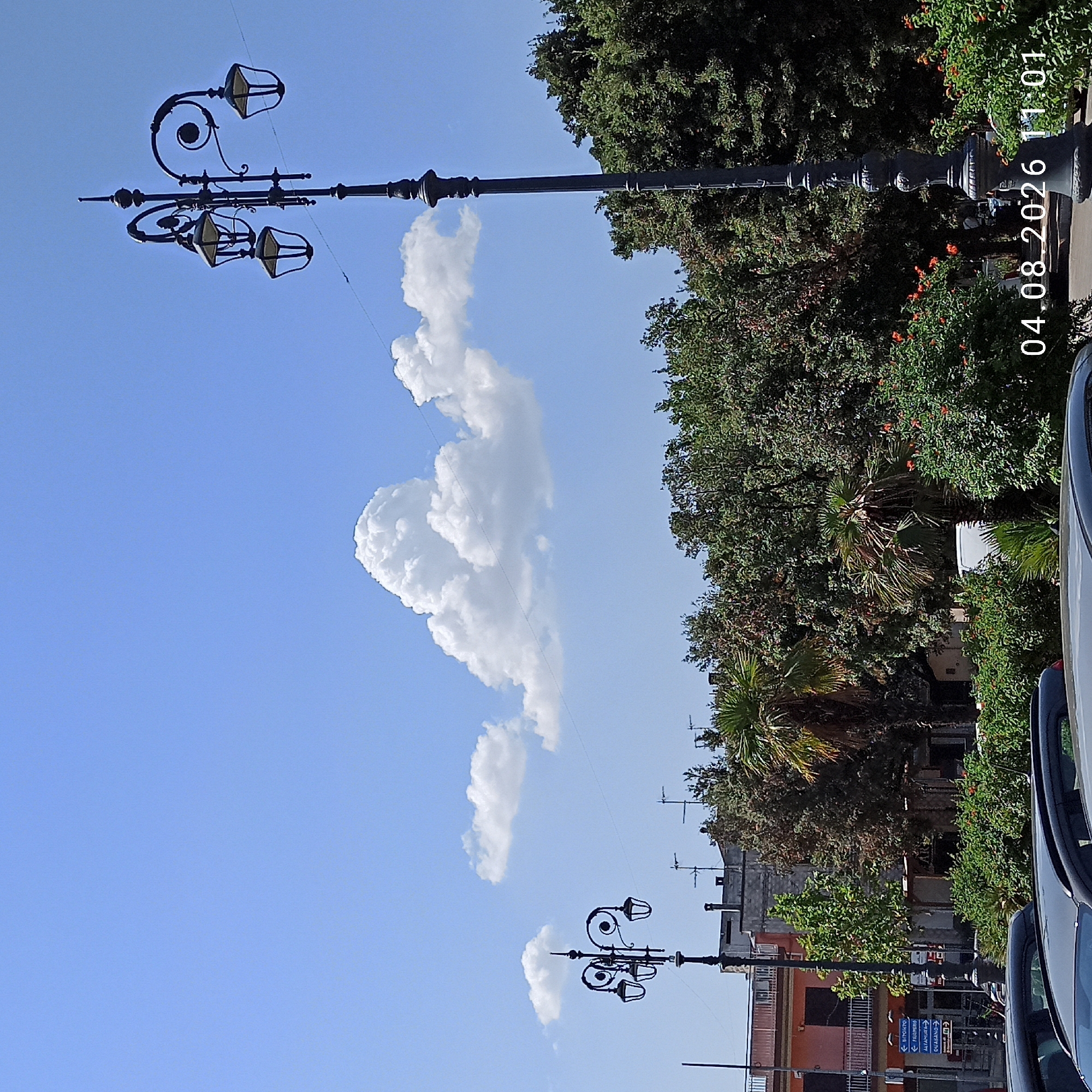 The image captures a daytime scene in Bitonto, Italy, with a clear blue sky and scattered white clouds. In the foreground, parts of two dark-colored cars are visible. A street lamp with an ornate, dark metal design stands tall on the right side of the image. To the left, another similar street lamp is visible. Behind the lamps and cars, there is lush green foliage and trees, with some buildings peeking through. A building with a salmon-colored facade and dark windows is on the far left. A road sign with Italian text is also visible near the left street lamp. The date and time "04.08.2026 11:01" are superimposed on the lower right corner of the image. The overall impression is of a street or public square in a city during daylight hours.