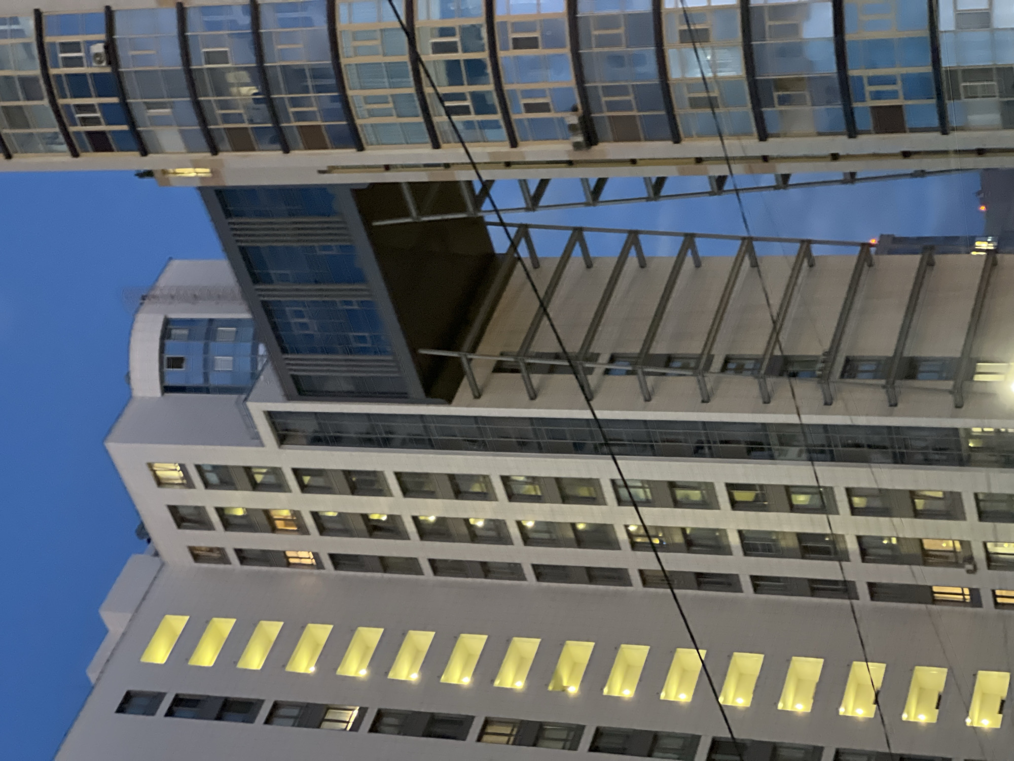 The image shows a contemporary multi-story residential building at dusk, with a clear blue sky above. The building features a light-colored facade with numerous windows, some of which are illuminated with a warm yellow light, suggesting interior lighting. A prominent architectural feature is a glass-enclosed walkway connecting two sections of the building, supported by a metal structure. To the right, another building with a facade of blue-tinted windows is visible. The setting appears to be urban, with electrical wires crossing the frame, hinting at power distribution infrastructure. The location is identified as Dachnoe, Russia. There are no people or animals visible in the frame. The overall scene conveys a sense of modern residential architecture in a twilight urban environment.
