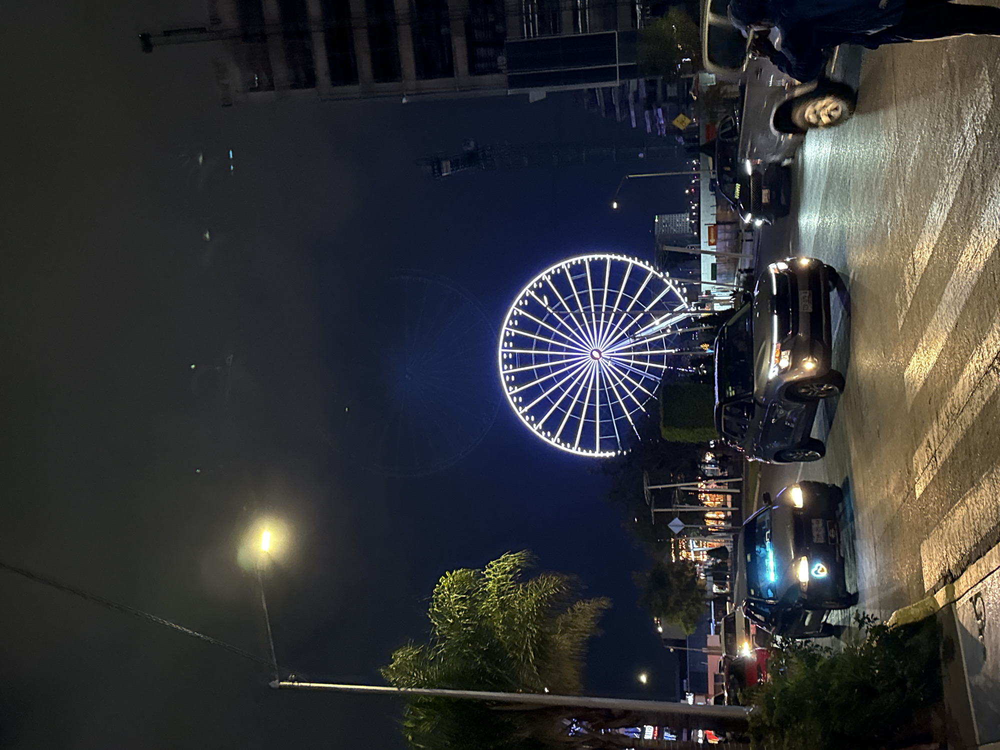 The image shows a street scene at night in Puebla, Mexico. A large Ferris wheel is illuminated in the background. Cars are parked and driving along the street. A person is visible standing near the cars, partially obscured. Streetlights illuminate the area, and buildings are visible in the background. Vegetation is present along the side of the road.