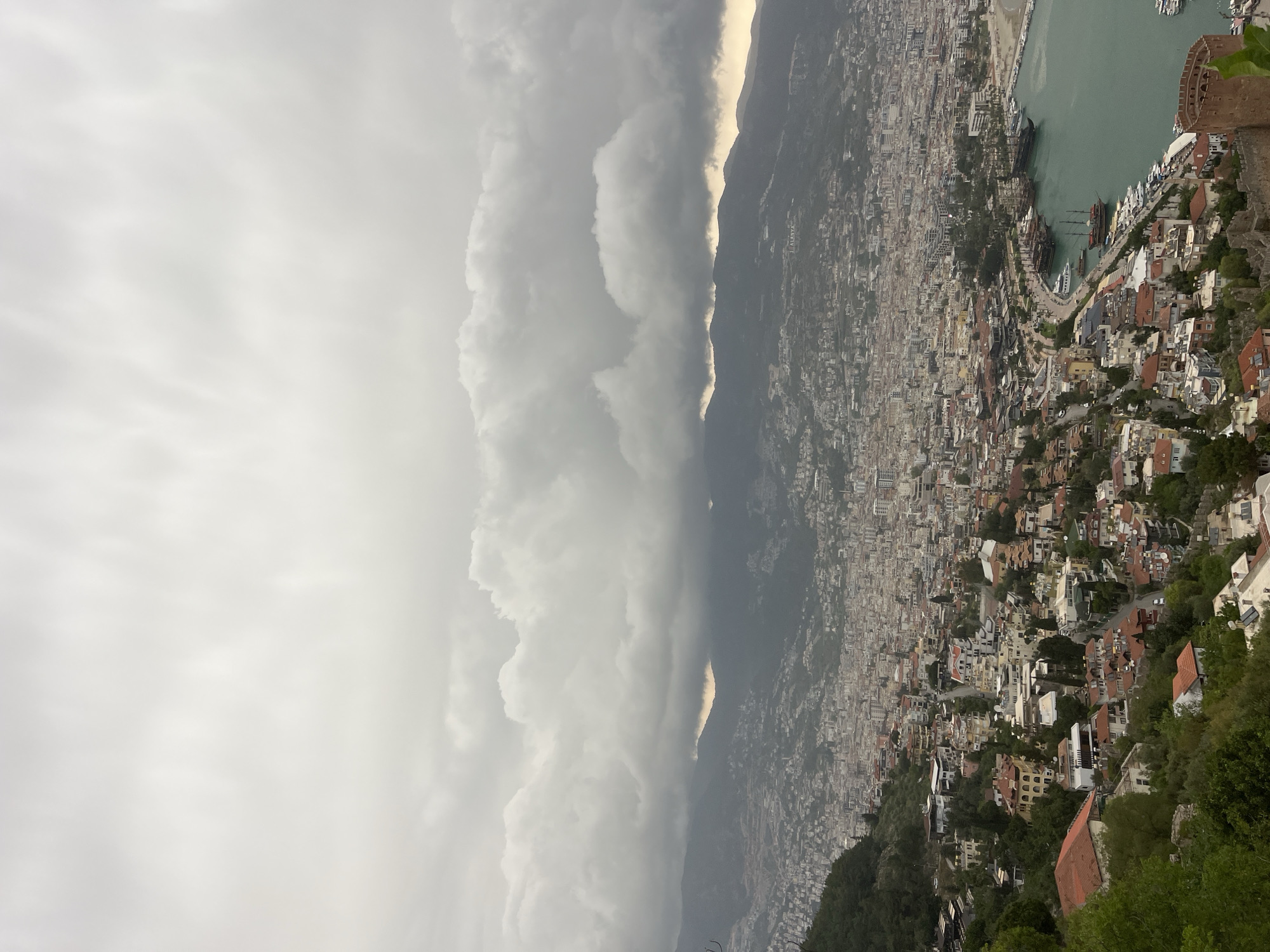 This panoramic photograph, presented in a portrait orientation but capturing a sweeping landscape, showcases a coastal city under a dramatic, cloudy sky. The left side of the image is dominated by an expansive, overcast sky filled with large, grey cumulus clouds that stretch across the horizon. Some brighter patches of cloud are visible, particularly near the top and along the mountain ridge, hinting at diffused light beyond the dense cloud cover.
A dark, undulating mountain range runs horizontally across the middle of the frame, partially obscured by the heavy cloud base and atmospheric haze. To the right of the mountains, a vast cityscape spreads out, densely packed with buildings that extend from the mountain slopes down to the coast. The architecture primarily consists of low-rise structures with a mix of light-colored facades and reddish-brown terracotta roofs. Green foliage and trees are visible, dotting the urban landscape and clinging to the hillsides.
Further to the right, a tranquil, turquoise-green body of water, likely the Mediterranean Sea, comes into view, forming a curving harbor. Several large, traditional wooden tour boats or galleons are moored along the waterfront, alongside smaller vessels. A distinctive reddish-brown stone tower or fortress wall, identifiable as part of Alanya Castle, stands prominently on the far right, overlooking the harbor and city. The overall atmosphere is moody due to the overcast weather. The scene clearly depicts Alanya, Turkey. No people or visible text are present.