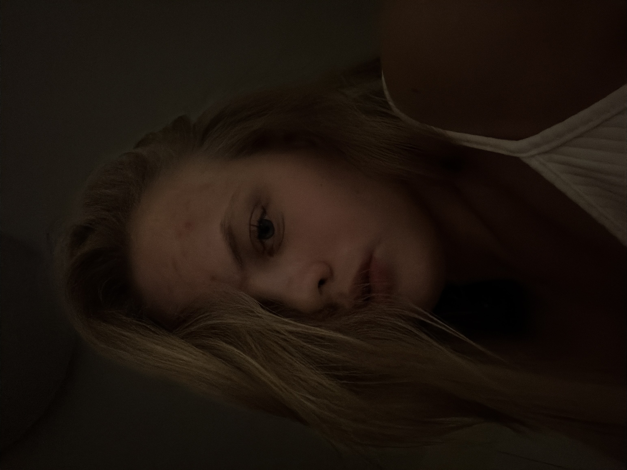 A fair-skinned young woman with blonde hair lies on her side in a dimly lit room. Her head is tilted towards the viewer, and some of her hair obscures part of her face. She wears a white tank top. The background is dark and indistinct. The location is not discernible from the image.