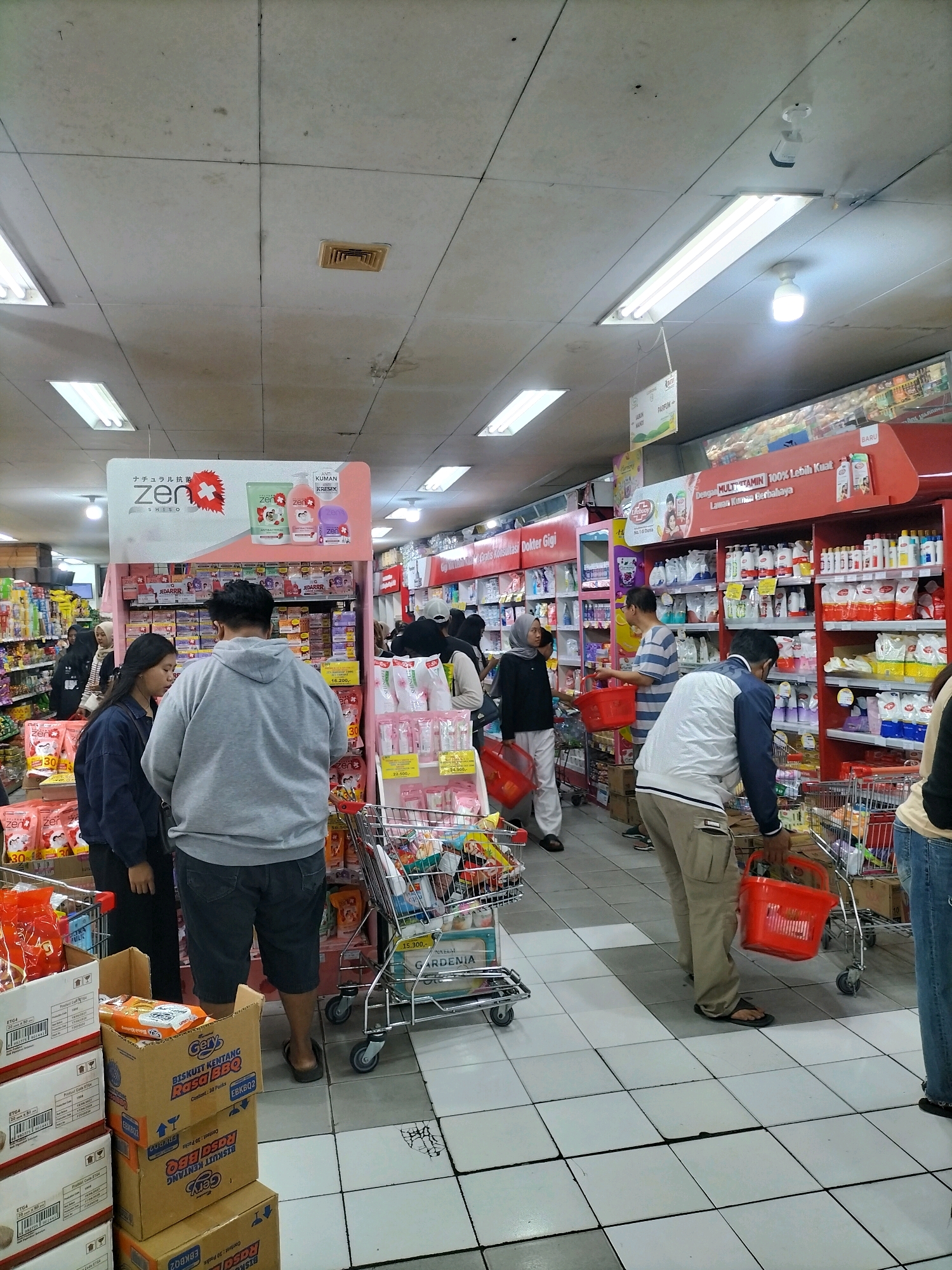 This is a full shot of the interior of a supermarket in Malang, Indonesia, as indicated by the Indonesian language on product packaging and signage. The scene is brightly lit by overhead fluorescent lights and appears to be a busy shopping day.
Several people are engaged in shopping activities. In the foreground, a couple stands facing a display of personal care products. The woman on the left is wearing a dark blue jacket and black pants, while the man in the center is in a grey hoodie and dark shorts. They appear to be browsing.
Further into the aisle, a man in a striped shirt holds a red shopping basket and is looking at products on a shelf. To his right, a man in a white jacket and light brown pants pushes a shopping cart, carrying a red basket. Another person in a black shirt and jeans is visible on the far right, also near a shopping cart. Other shoppers are visible in the background, dispersed among the aisles.
The environment is filled with merchandise displayed on shelves and stacked in boxes. Shelves are stocked with various consumer goods, including what appears to be toiletries, cleaning supplies, and food items. Promotional signage is visible, including a large pink display featuring "Zen Shiso" products at the front. Shopping carts and baskets are common objects throughout the scene. The floor is tiled with a light-colored grid pattern. The overall impression is that of a typical retail environment within a busy Indonesian supermarket.