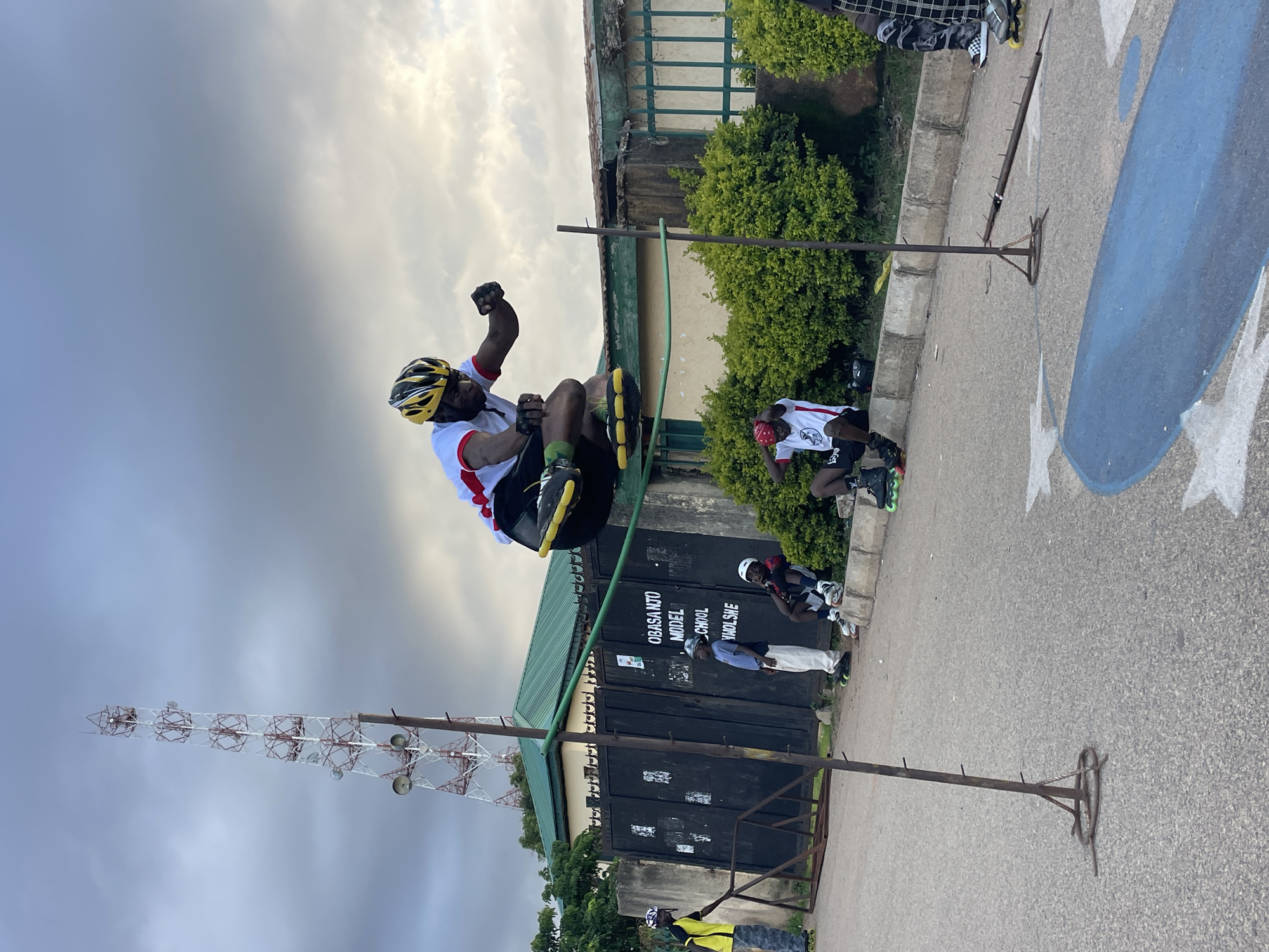 In Jos, Nigeria, a man on rollerblades performs a jump over a horizontal bar set between two poles. He is wearing a yellow helmet and a white and red shirt. The jump is taking place on a paved road in front of a building with a dark gate that reads "OBASANJO MODEL SCHOOL WADLSHIE." Several other people are present, some sitting on a curb and others standing, also wearing rollerblades and helmets. A tall communication tower is visible in the background, and the sky is overcast with clouds. Lush green bushes line the area behind the curb. The overall scene suggests an informal gathering or practice session for rollerblading stunts.