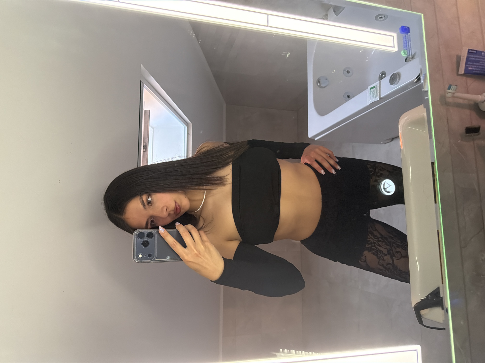 A young woman in a black crop top and leggings stands in a bathroom, taking a mirror selfie. The bathroom appears modern, with a large mirror, a bathtub, and typical bathroom fixtures. The location cannot be determined as Gabrovo, Bulgaria with the provided image information.
