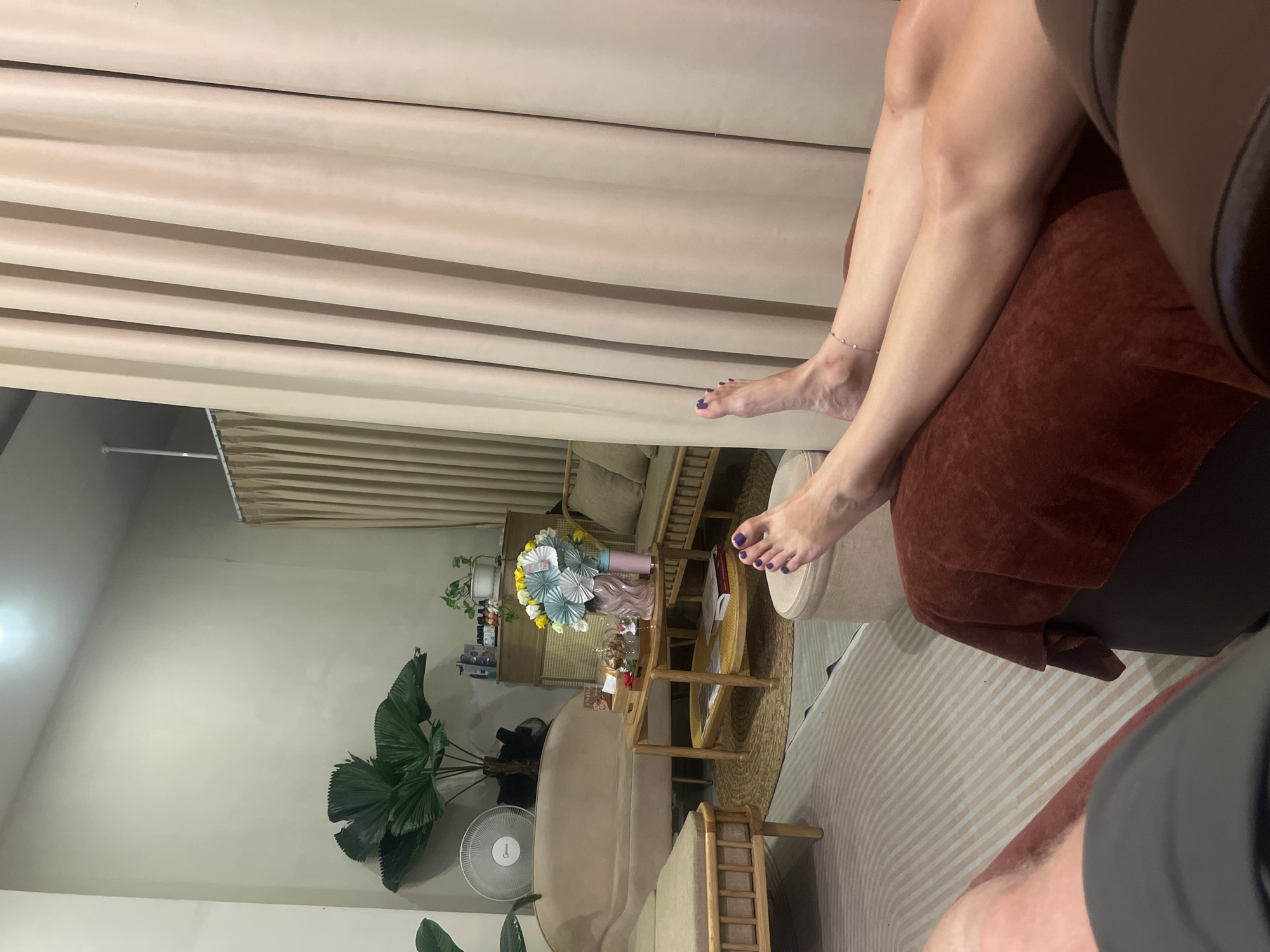 The image shows an interior scene in an unknown location. There are legs and feet resting on an upholstered surface, possibly a chair or couch. The feet are painted with purple nail polish, and one leg has an anklet. A set of curtains are visible in the background, and there is a seating area with various pieces of furniture, including a couch, table, and decorative items. A large potted plant and a fan are also in view.