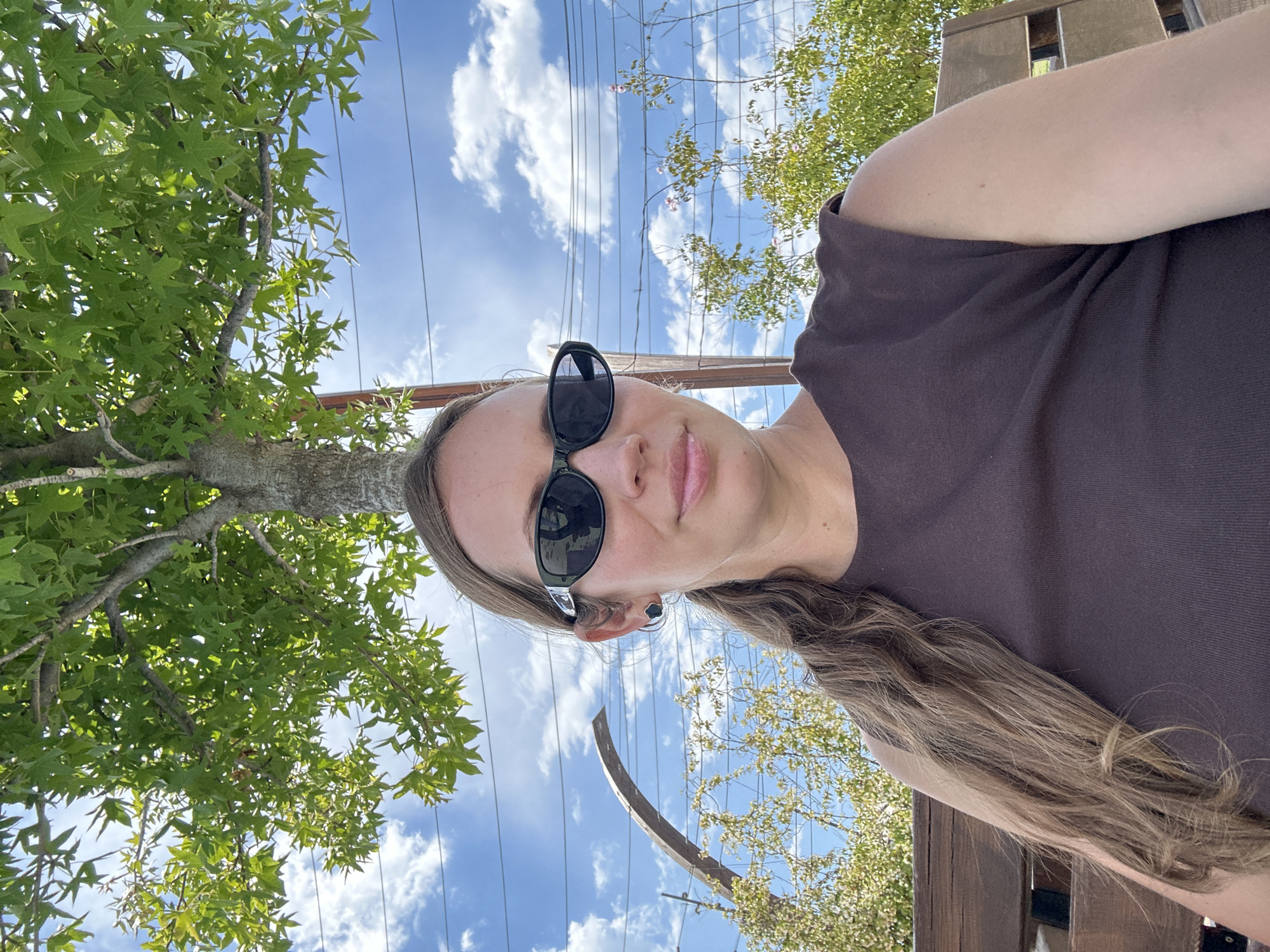 A woman with long brown hair, wearing black sunglasses and a brown top, is looking at the camera. She is standing outside on a sunny day with some trees and a blue sky with clouds behind her. There are power lines in the background. A wooden structure is visible on the right. The location is likely in Fethiye, Turkey, but this cannot be verified from the image.