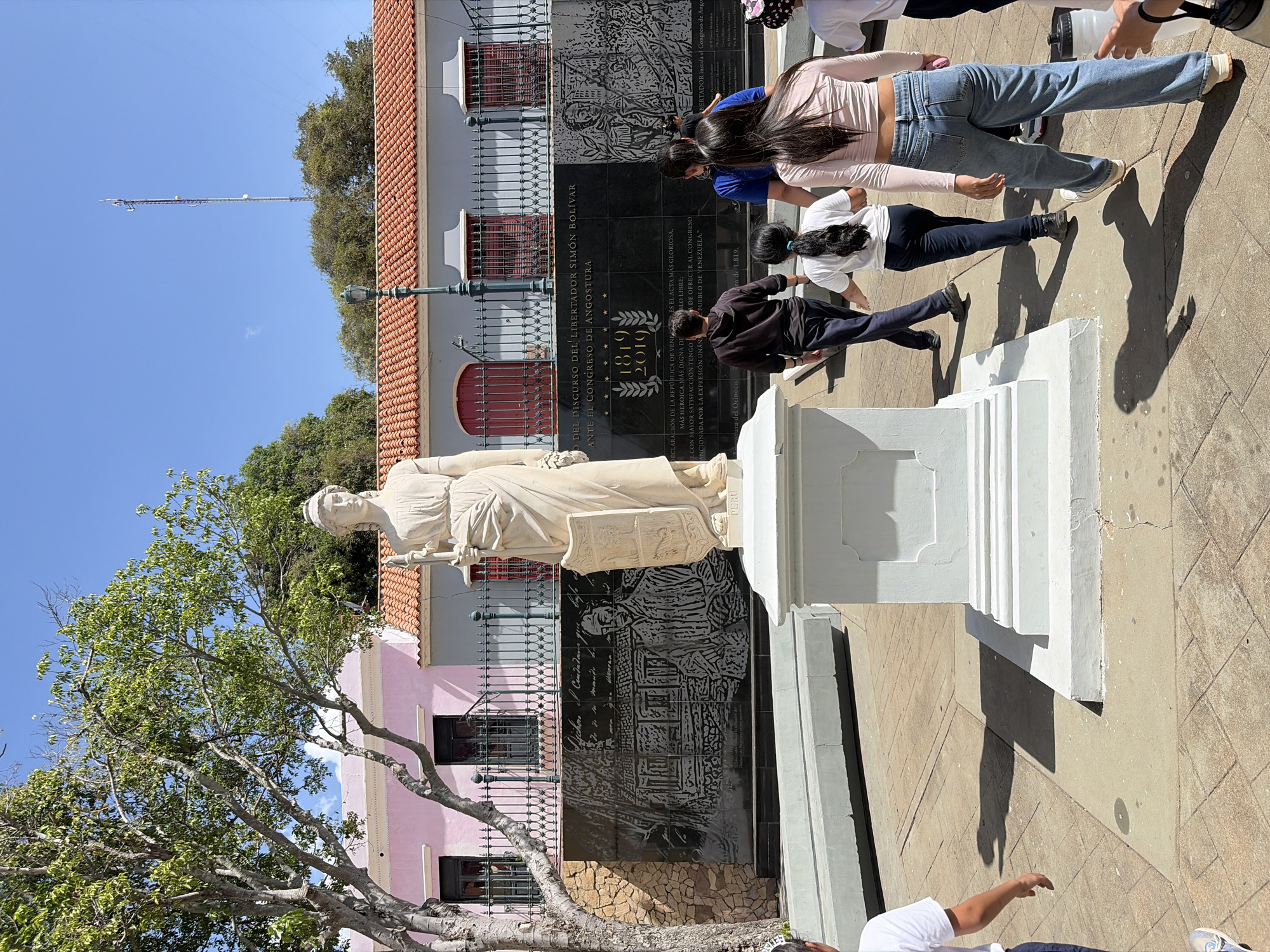The image shows a statue of Simon Bolivar standing in front of a black monument, likely in Ciudad Bolivar, Venezuela. The monument includes the years "1819" and "2010," and the text appears to be a declaration of the Republic of Venezuela. The statue is on a white pedestal. Several people are standing around the monument, appearing to observe or interact with it. A tree is visible to the left of the statue, and several buildings can be seen in the background. The sky is blue, and the scene appears to be outdoors in daylight.