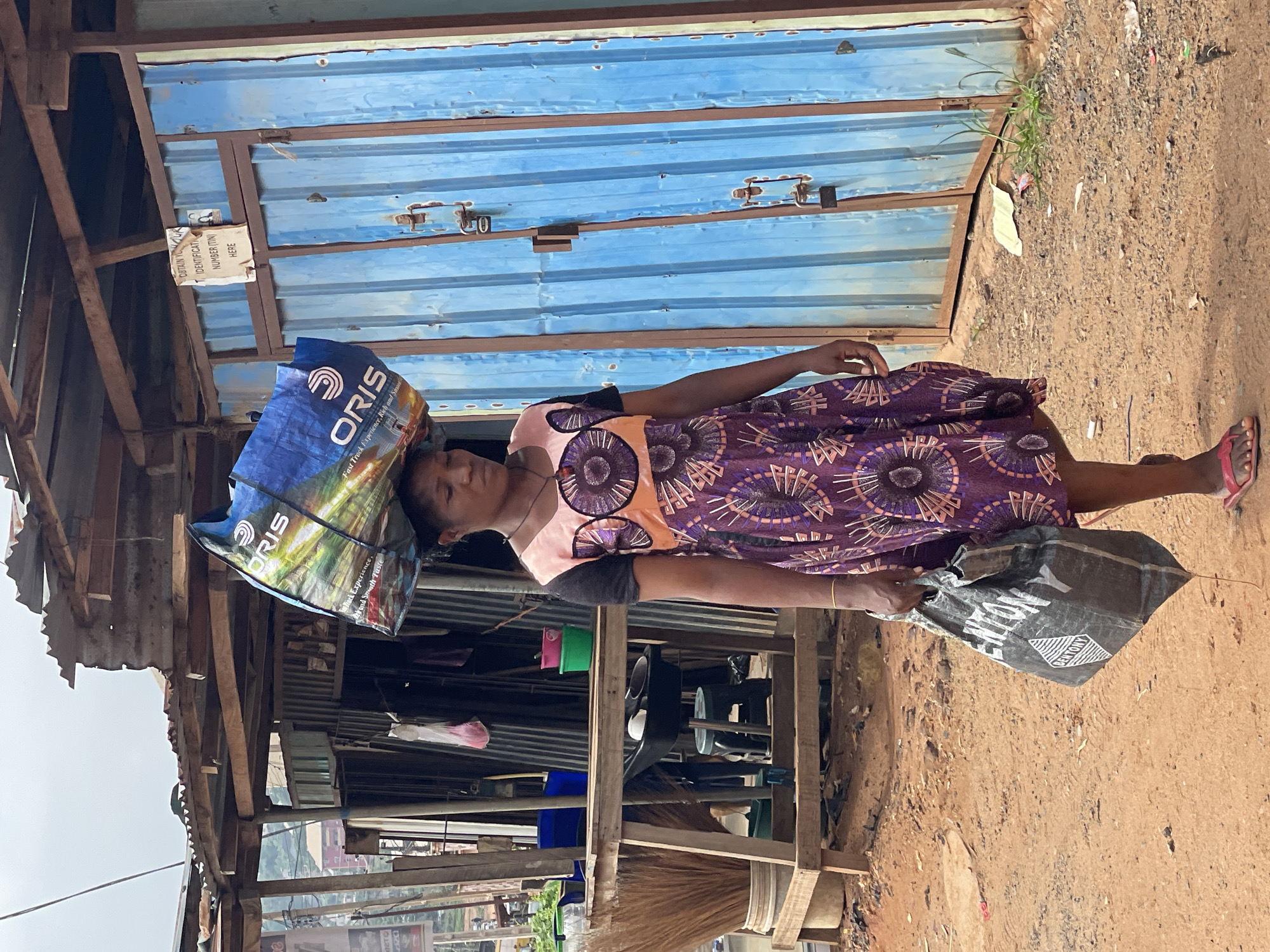 The image captures a woman in Nkpor, Nigeria, carrying items both on her head and in her hand, standing beside a corrugated metal structure. The woman is wearing a dress with a purple and brown circular pattern, and sandals on her feet. On her head, she balances a blue bag with the word "ORIS" printed on it. In her hand, she carries a black bag.
The corrugated metal structure is painted blue and appears to be a shop or storage area. A sign is posted above a bag on her head, displaying text, including the word "IDENTIFICATION". The ground is dirt. The background features additional structures and greenery, suggesting an urban environment.