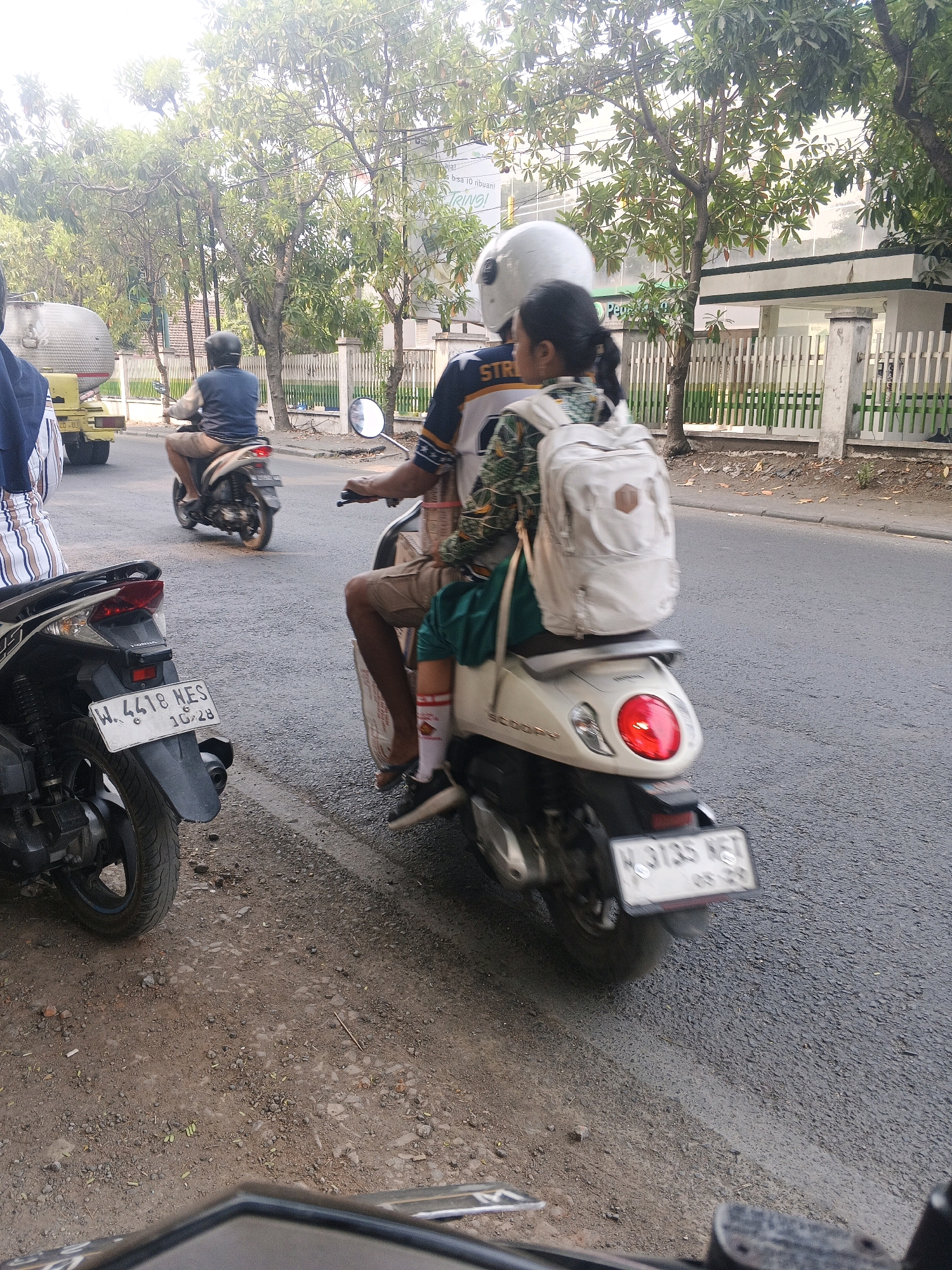 In Krian, Indonesia, a group of motorcyclists navigates a paved road. A man wearing a blue shirt and brown shorts rides a white scooter, while a woman with a white backpack and a green scarf rides behind a man wearing a striped shirt and brown shorts on another scooter. To the left, a dark-colored scooter is parked on a dirt shoulder. In the background, trees line a white fence, and a yellow truck with a cylindrical tank is visible. The sky is bright, indicating daylight.