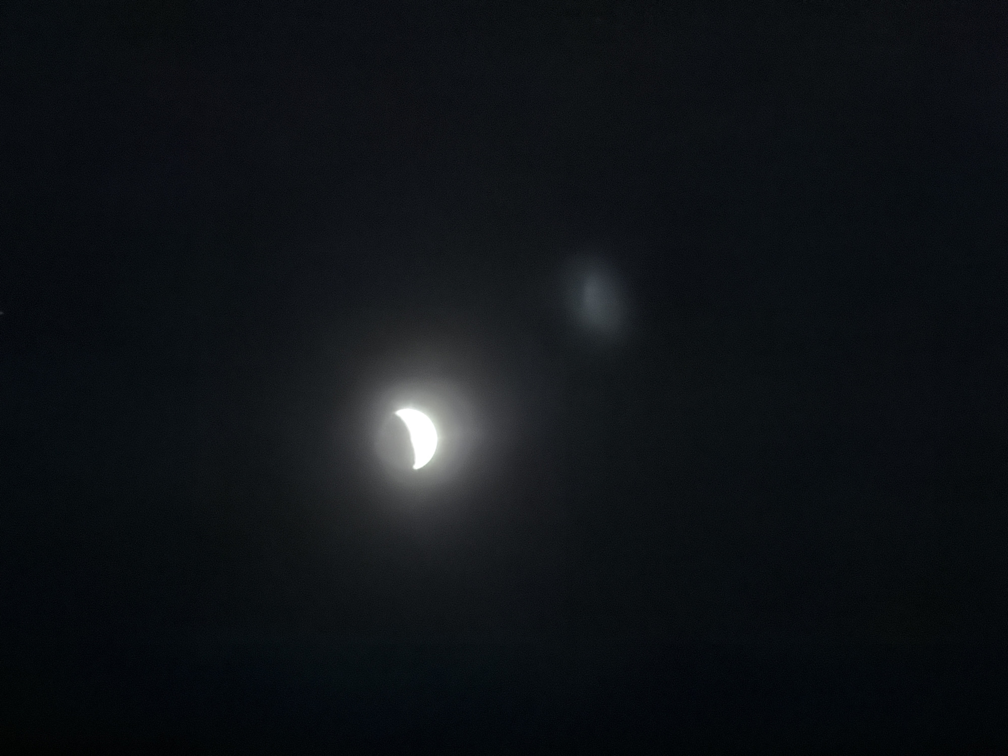 The image depicts a partial solar eclipse against a dark sky. The eclipsed sun appears as a bright crescent shape. There's a blurry patch of light to the right of the eclipsed sun. The photo was taken at night or under very low light conditions, and there is no visible indication of a specific location within Paris, France. No people, objects, or actions besides the eclipse are discernible.