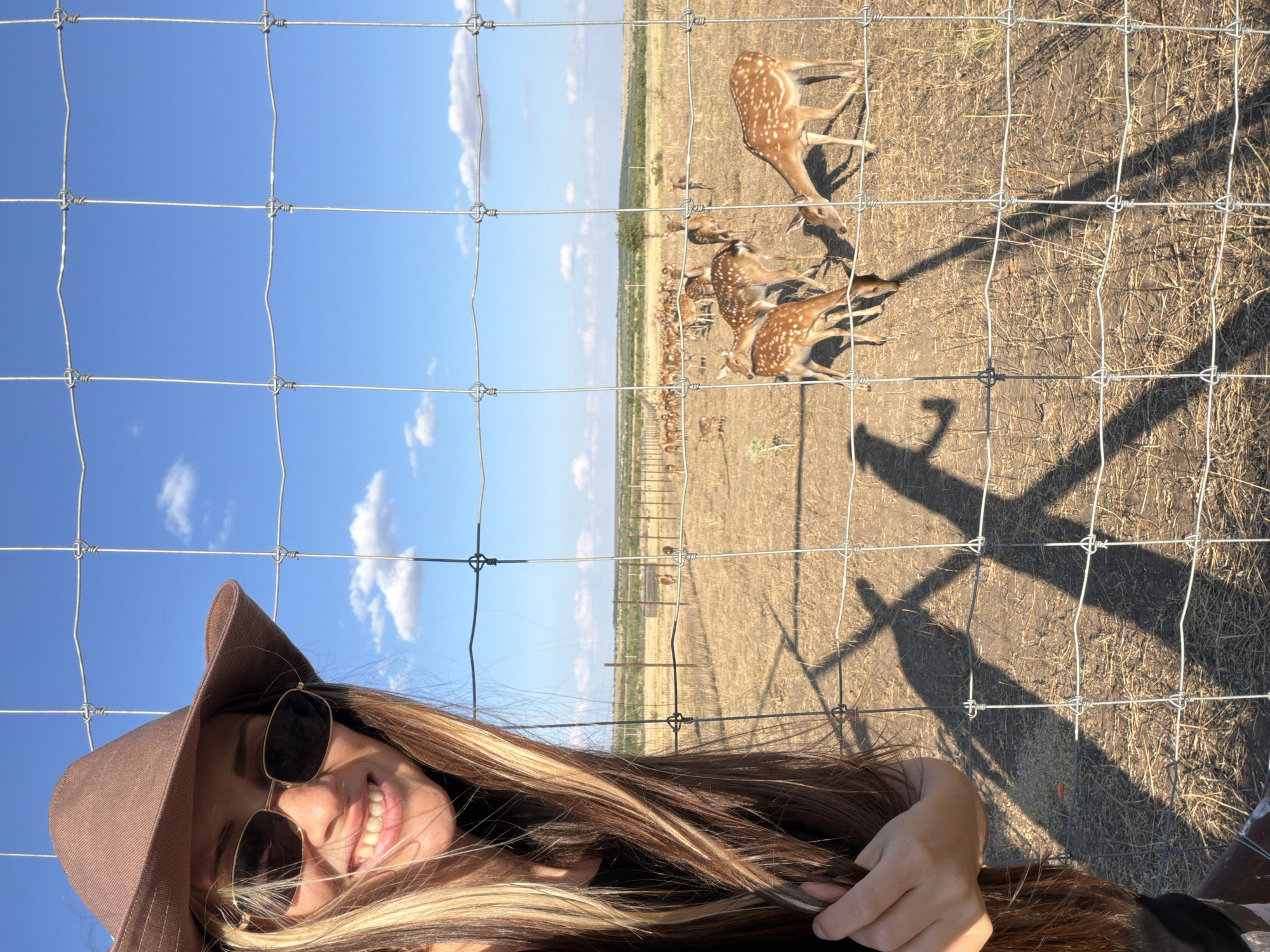 A woman wearing a brown hat and sunglasses smiles at the camera, her long brown and blonde hair cascading over her shoulders. She is standing behind a wire fence, with a herd of spotted deer visible in the dry, grassy field beyond. The sky is blue with scattered white clouds. The environment appears to be an open, rural area, possibly a wildlife preserve or ranch. The shadows of the woman and the fence are cast on the ground, indicating a sunny day. The location is identified as Gorny Balykley, Russia.