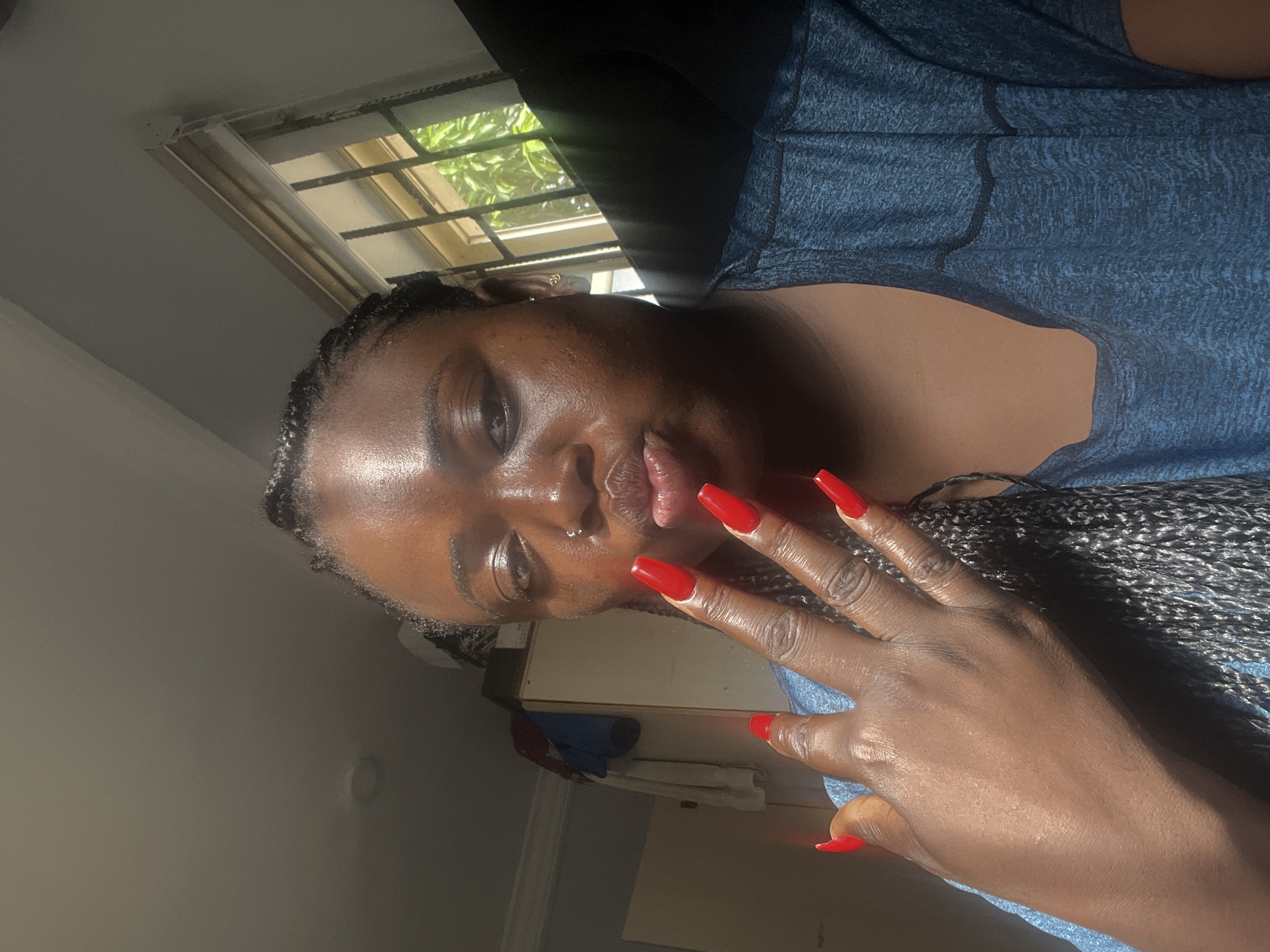 A close-up portrait shows a Black woman indoors, captured from a rotated perspective. She looks directly at the camera with a neutral, posed expression, her lips slightly parted. Her hair is styled in braids, and she wears a small silver nose ring. Her left hand is raised in the foreground, showcasing long, sharp, bright red nails. She is dressed in a blue short-sleeved top with a subtle textured pattern.
Strong, natural sunlight enters from a window positioned in the upper part of the frame, brightly illuminating her face and hand and creating highlights on her skin. Through the window, lush green foliage is visible, suggesting trees, and the window has horizontal security bars. The background consists of a plain wall to the left and indistinct indoor elements behind her arm. The bright lighting indicates it is daytime. No text is visible.