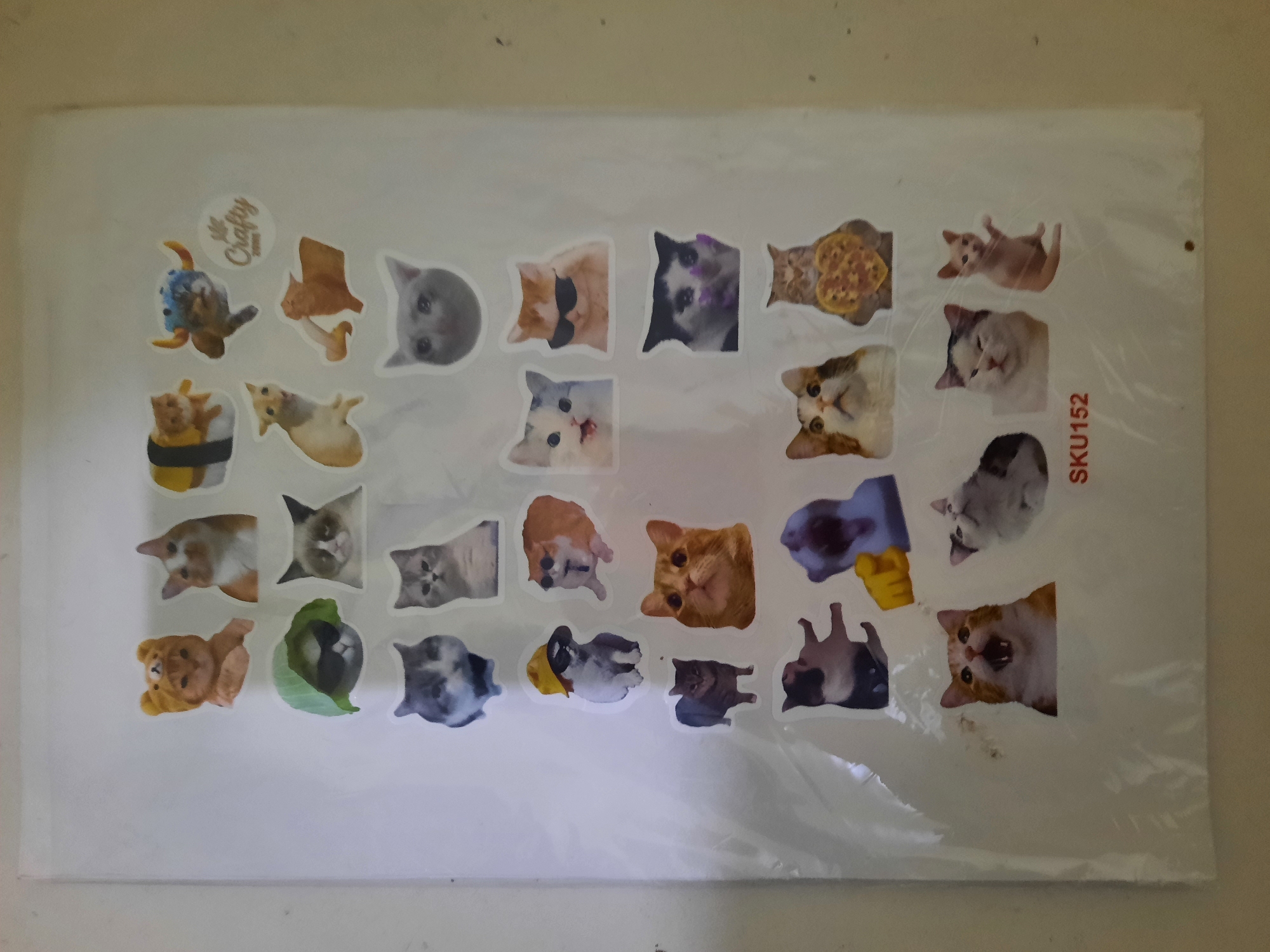 A sticker sheet featuring various cat illustrations is displayed against a light-colored, textured wall. The sticker sheet is vertically oriented and has a plastic protective covering. The cats are depicted in different poses and with various accessories, including hats, sunglasses, and costumes. Some notable cat illustrations include a cat in a yellow bear hat, a cat wearing sunglasses and a green leaf, a cat with a grumpy expression, a cat dressed as sushi, a cat with bull horns, a cat with a sad expression, a cat holding a heart-shaped object, and cats in various playful or surprised stances. The bottom of the sticker sheet has "SKU152" printed on it. The image is captured from a slightly elevated angle. The lighting is even, with no harsh shadows. The scene appears to be indoors. There is no discernible connection to Lhokseumawe, Indonesia.