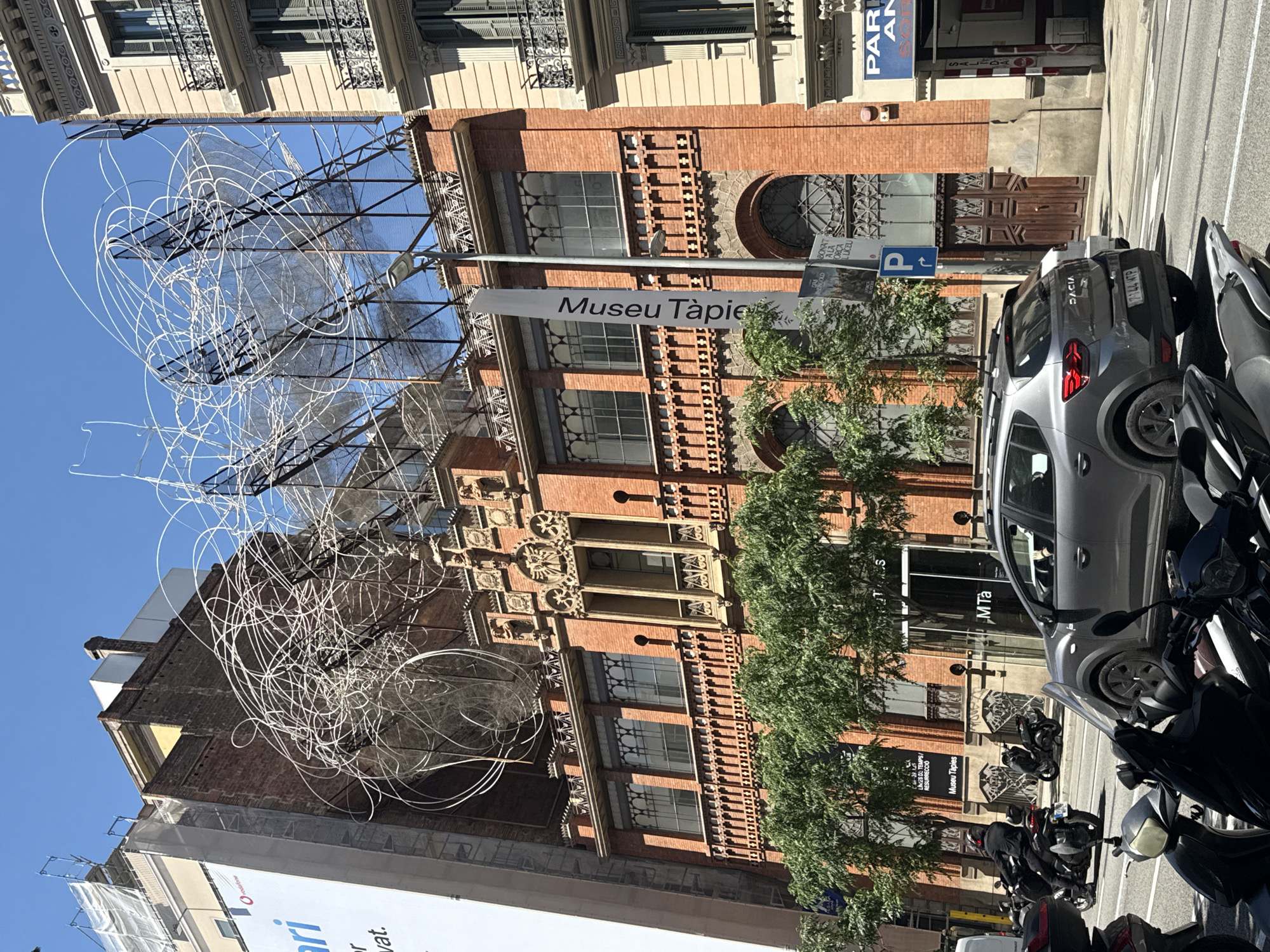 The image shows a street view in Barcelona, Spain, featuring the Fundació Antoni Tàpies. The museum building is constructed with red bricks and has intricate architectural details. A contemporary art sculpture composed of metal wire is installed on the building's rooftop. In front of the building is a street with parked cars, including a gray Dacia, and numerous scooters. A street sign indicates the name of the museum: "Museu Tàpies". Advertising banners from Vodafone are also visible. The sky is clear and blue.