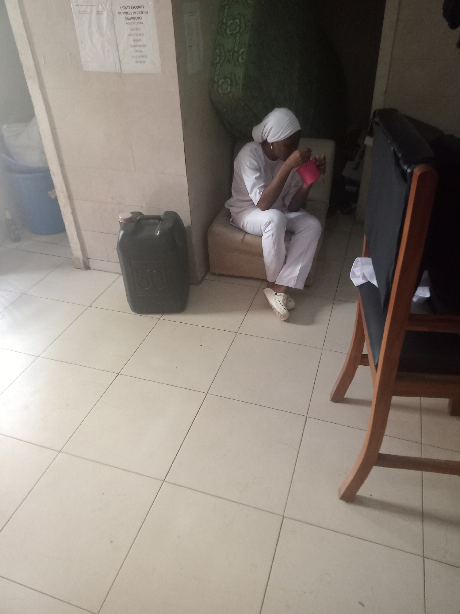 The image captures an indoor scene with a woman in white scrubs and a head covering, possibly a nurse or healthcare worker, sitting on a light-colored cushioned seat and eating from a pink container. Beside her is a large black plastic container labeled "50" with a red cap. Adjacent to the container is a white wall with a notice titled "ESTATE SECURITY NUMBERS IN CASE OF EMERGENCY", listing various contact numbers. To the right, a wooden chair with a black seat is partially visible. The scene appears to be set on a tiled floor in a hallway or waiting area. The location of Gwarinpa, Nigeria, is deduced from the listed contact numbers on the notice, which are Nigerian phone numbers.