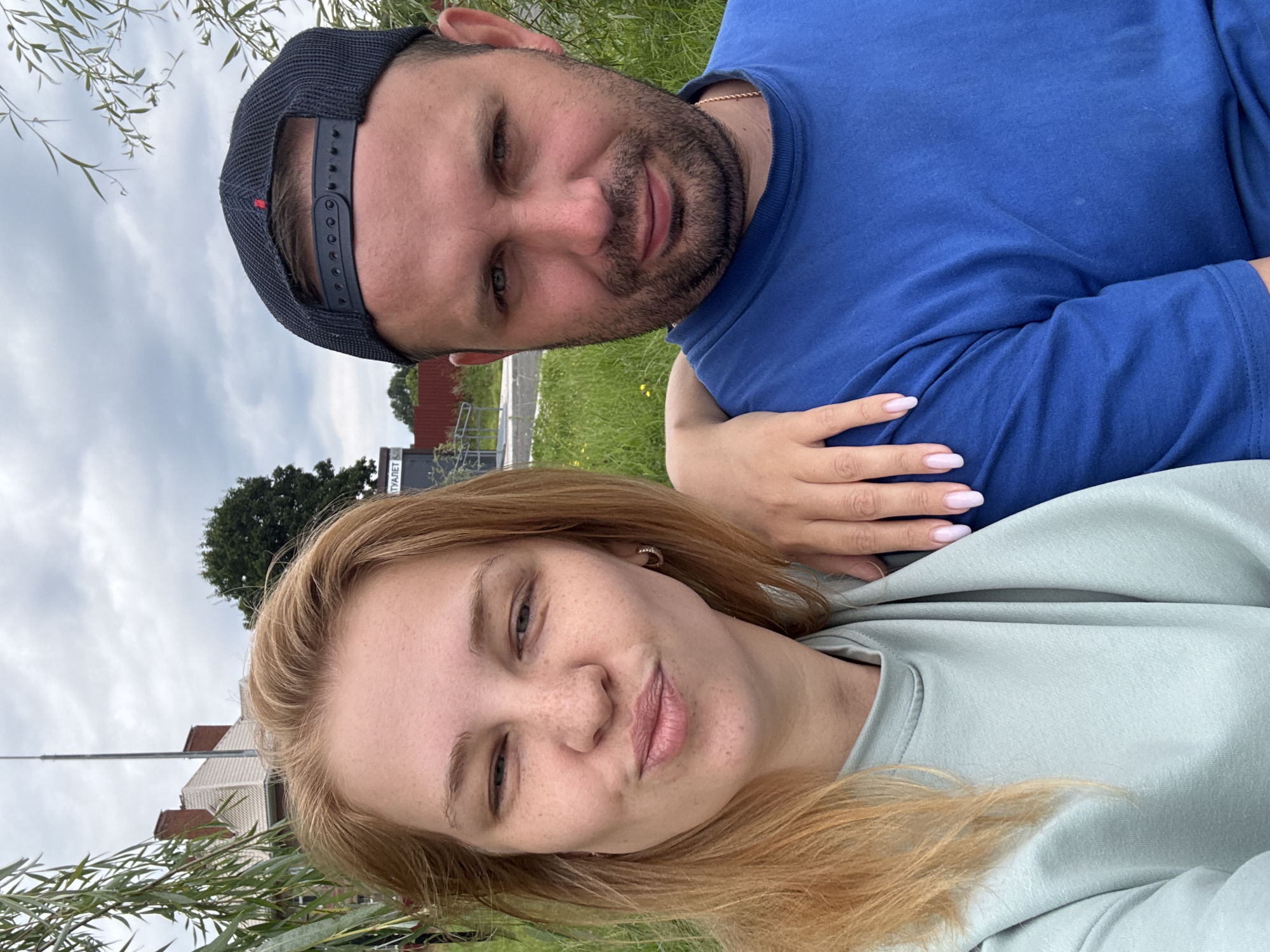 In Breytovo, Russia, a close-up, waist-up selfie captures a couple outdoors under a cloudy sky. The woman, positioned on the left, has light blonde hair and is wearing a light teal sweatshirt. She is making a puckered-lip expression, looking towards the camera, with her left hand resting on the man's shoulder. Her right arm is not visible. She has light skin with freckles and is wearing small hoop earrings.
The man, on the right, is wearing a blue t-shirt and a dark baseball cap. He has short, dark hair and stubble. He is looking directly at the camera with a neutral expression. A gold chain necklace is visible around his neck. His left arm is around the woman's shoulder, with his hand resting on her back.
Behind the couple, a building with a red-tiled roof and a brick chimney is partially visible to the left, with some greenery and trees in the background. A sign with Russian text is visible on a pole to the right of the building. The ground is covered in grass.