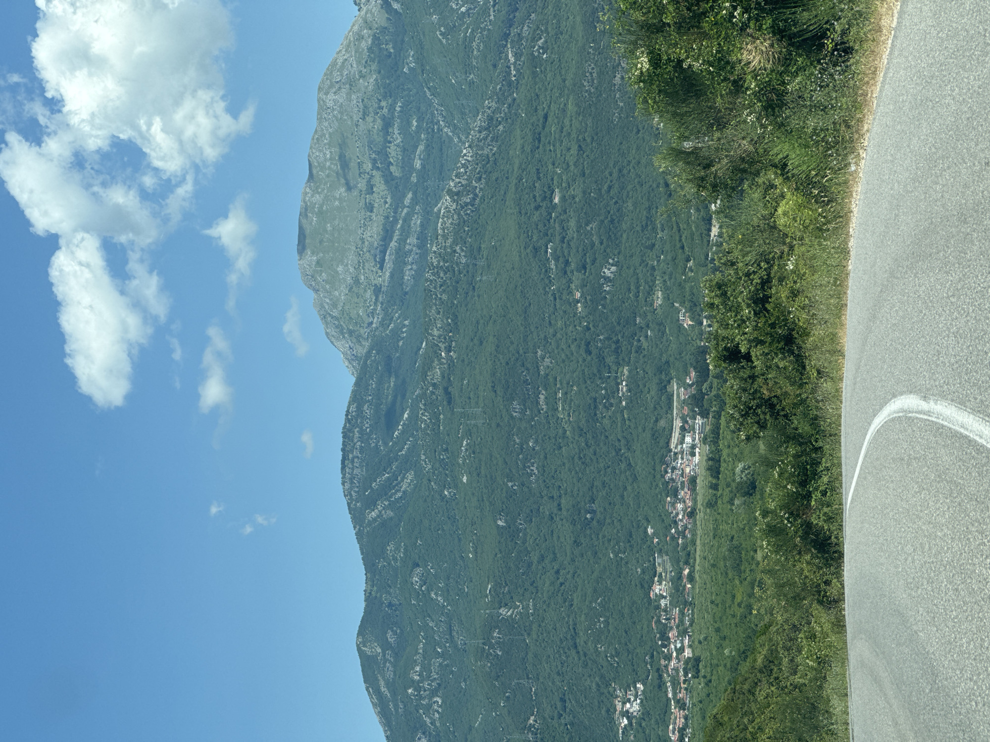 The image presents a sunlit, panoramic landscape in Kubasi, Montenegro. In the immediate foreground, an asphalt road curves to the left, marked by a solid white line along its inner edge. The right side of the road is bordered by dense green vegetation, consisting of shrubs, grasses, and some plants with small white flowers.
The midground features a verdant valley, heavily forested with trees and bushes. Nestled within this valley, a village is visible, characterized by numerous houses with predominantly light-colored roofs, scattered across the lower slopes. Faint power line structures or utility poles are discernible extending across the tree-covered mountainside above the village.
The background is dominated by large, imposing mountains, richly covered in green foliage, with some sections revealing rocky outcrops. The peak of the highest mountain is visible against the sky. The sky above is a clear, bright blue, accented by several white, fluffy cumulus clouds, primarily in the upper right quadrant of the frame. The overall lighting suggests bright daylight conditions. No individuals, animals, or vehicles are present in the scene.