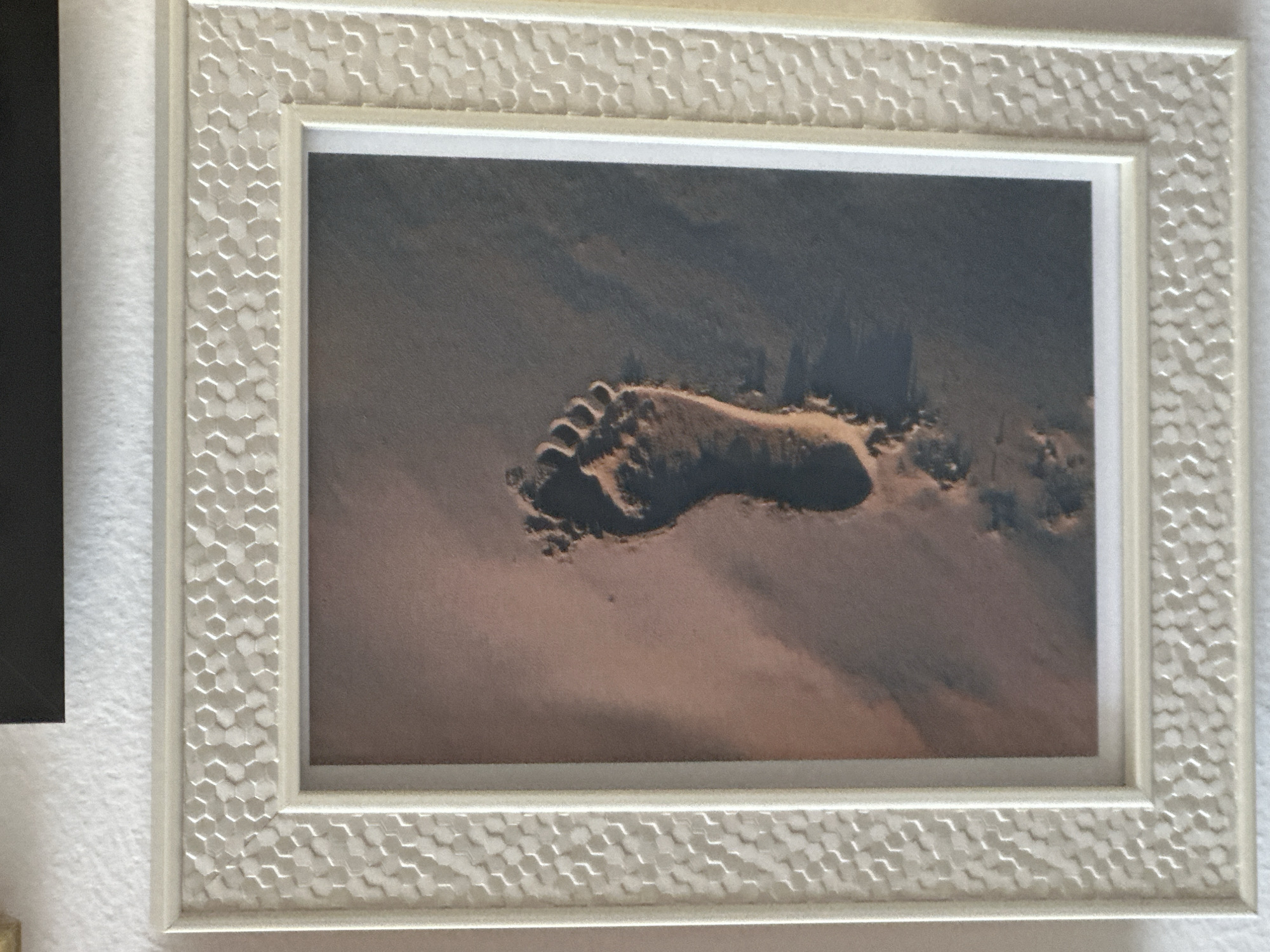 A framed photograph of a single footprint in the sand is displayed against a white wall. The frame is white with a textured honeycomb pattern around the outer edge. The photograph itself shows a footprint pressed into damp, reddish-brown sand. The heel of the footprint is visible on the right side of the image, with the toes pointing towards the left. There are also faint impressions of other footprints trailing away from the main one. The lighting suggests a shadow cast from the footprint, indicating a light source from the upper left. The overall impression is that of a moment captured in time on a beach.