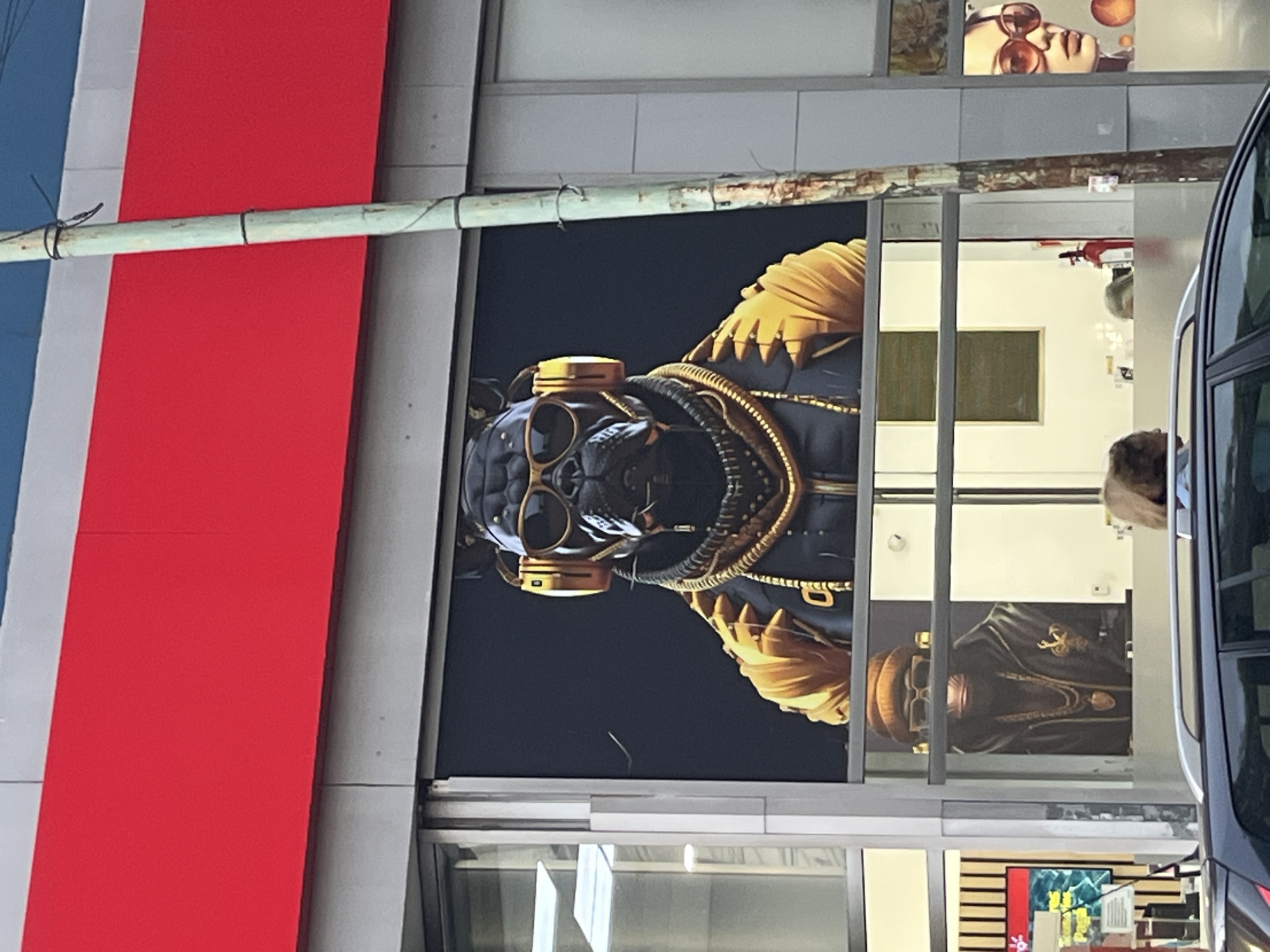The image, rotated 90 degrees clockwise, captures an urban street scene during daylight, centered on a building facade. A large, vertically oriented artwork is prominently displayed on a glass storefront. This artwork features a stylized, dark-colored animal head, resembling a bulldog, wearing golden-rimmed goggles and golden headphones or ear coverings. Its body is depicted in black and gold, with intricate details like a bandolier-style strap or chains across the chest and layered golden shoulder pads. The background of the artwork is a dark blue or black.
To the left of the storefront, part of a structure with vertical red, white, and blue panels is visible. A light-colored pole runs horizontally across the red and white sections, secured by black ties. The glass of the storefront reflects the interior of the building, revealing white walls and a dark rectangular object, possibly a screen or board. Above the main artwork, another smaller image or reflection shows a person's face wearing large sunglasses. On the far right, a portion of a dark-colored vehicle, likely a bus, is visible, along with a blurred reflection of a person's head. There is no visible text in the image. The scene portrays an artistic display in a commercial setting.