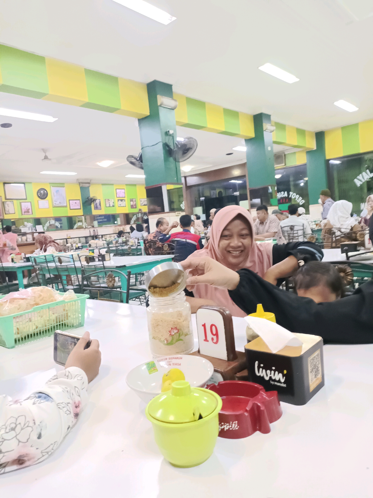 In the city of Dapur, Indonesia, a vibrant dining establishment features a bright green and yellow checkered ceiling. The interior is populated with diners seated at white tables. In the foreground, a person's hand, adorned with a floral-patterned sleeve, holds a smartphone and appears to be taking a photograph. On the table, a green basket contains crackers, a glass jar with a metal lid holds a granular substance, a white bowl holds yellow pieces, a lime green covered bowl, a red ashtray, and a tissue dispenser with the "Livin" logo. Behind this, a woman in a pink hijab smiles warmly. Other patrons are visible in the background, engaged in their meals and conversations within the bustling restaurant setting.