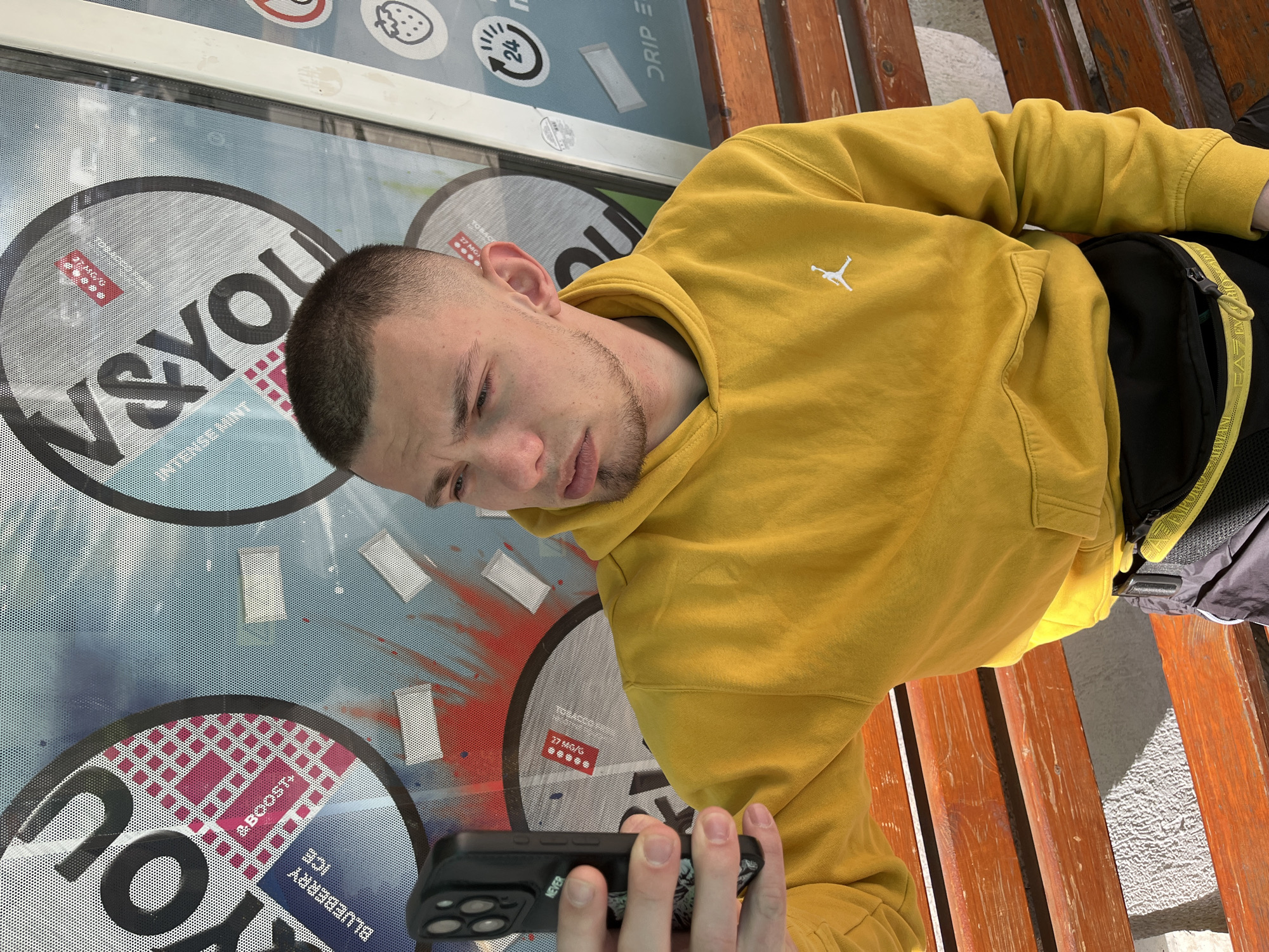 The image shows a young man with a buzzcut and a slight beard, wearing a yellow hooded sweatshirt with a white Jordan logo on the chest, looking at his smartphone. He is sitting on a wooden bench next to a glass display case featuring advertising for "W&YOU" nicotine pouches. The display includes flavors like "Intense Mint" and "Blueberry Ice", along with details like "27 MG/G", a "boost" graphic, a strawberry, and symbols for "24" hours and "Drip E". The man's right hand holds the smartphone. A black backpack is also visible to the right of the person. It is likely that the photo was taken in Burgas, Bulgaria, due to the brands of nicotine pouches advertised, as they are commonly available in that area.