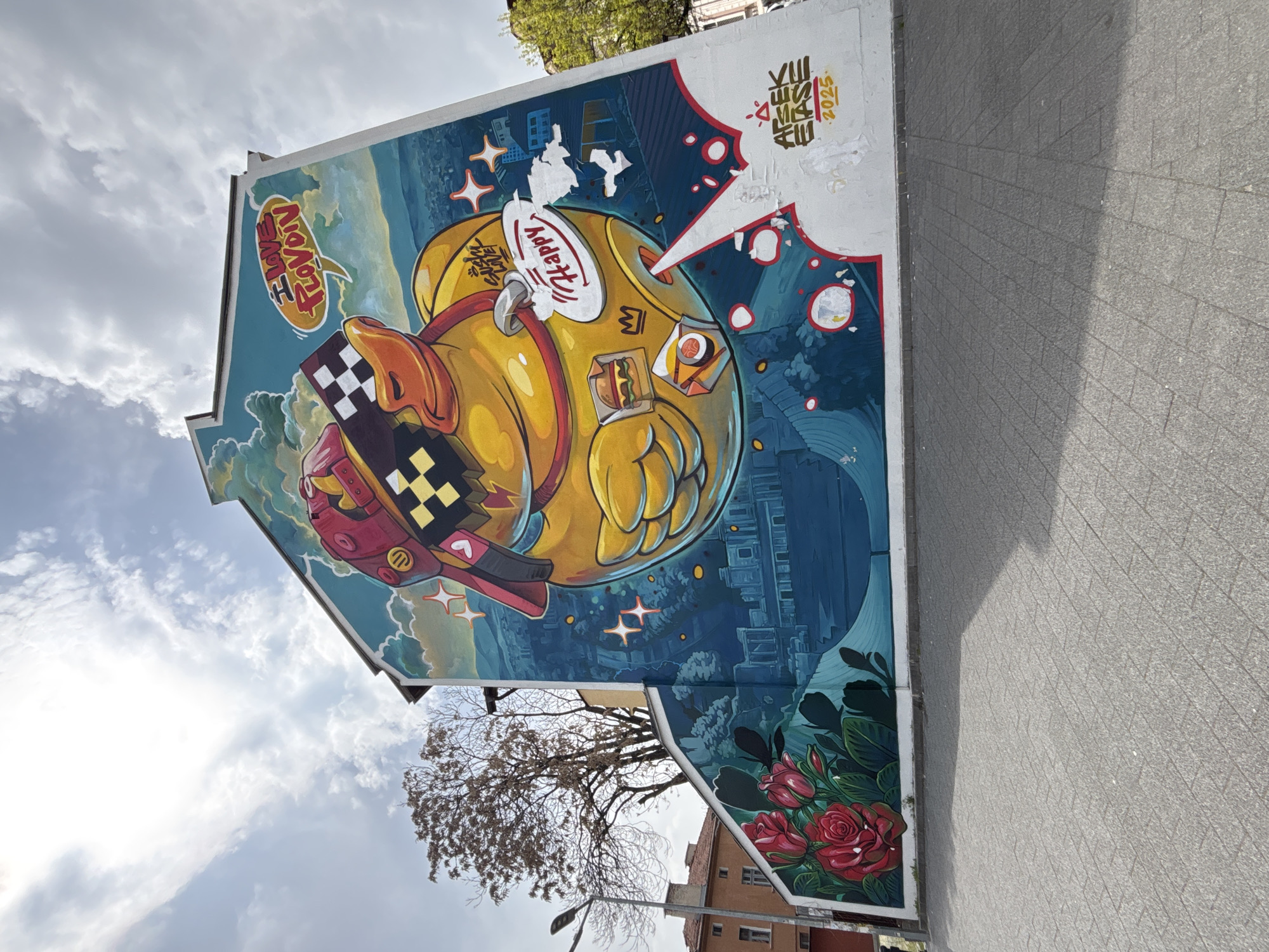 The image shows a mural on the side of a building. The mural features a stylized, yellow cartoon character resembling a duck or chick, wearing sunglasses with a checkered pattern. The character is holding food containers with a crown symbol on it. A speech bubble above the character says "I Love Plovdiv."
The background includes cityscapes and a stylized night scene. There is additional artwork in the upper right corner that includes text in a different language and the year "2025." Red roses are painted at the bottom of the mural. The building is adjacent to a paved area. A tree is visible in front of the building, with more buildings behind. The mural is in Plovdiv, Bulgaria.