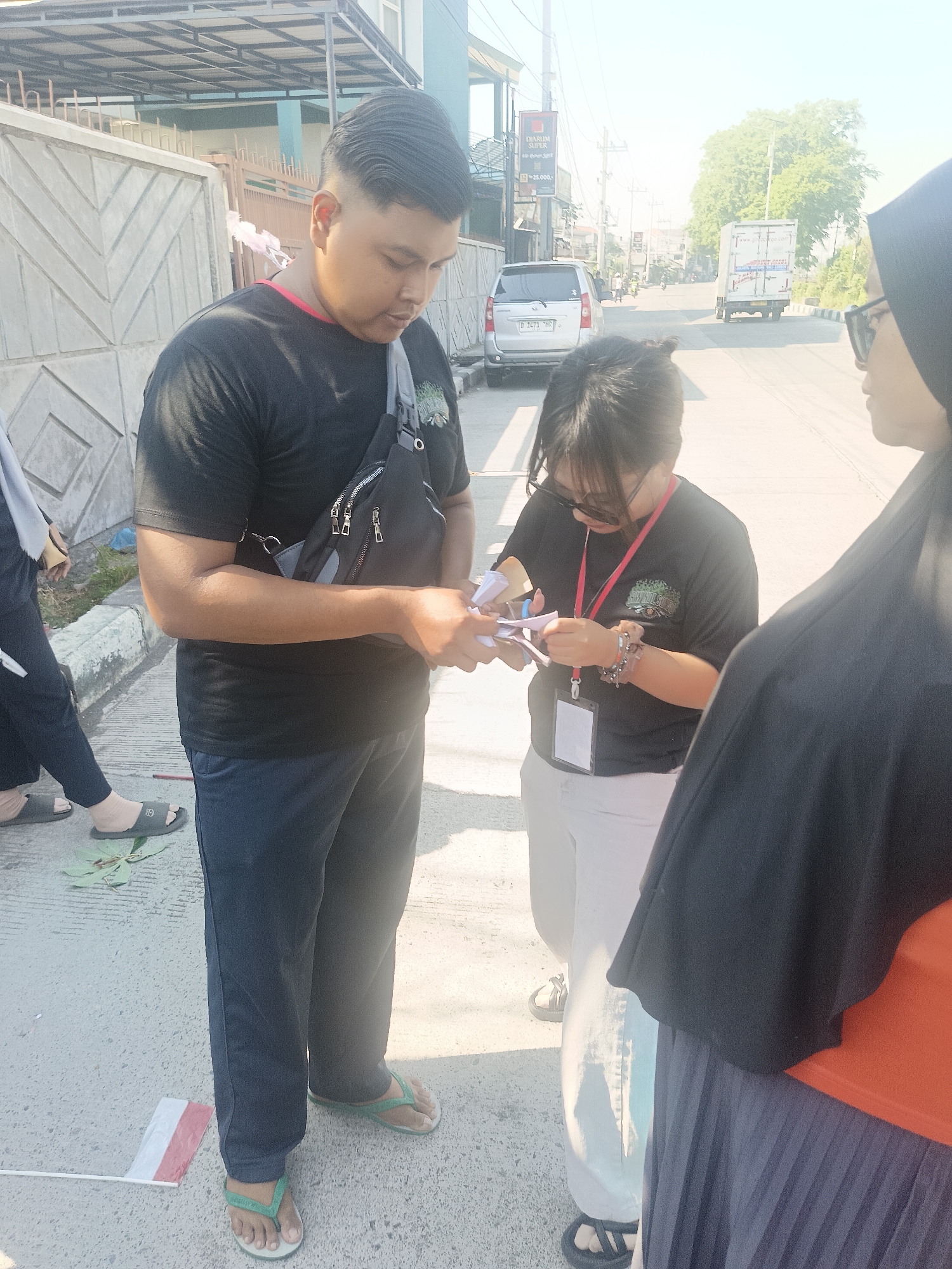 The image depicts an outdoor scene in Gedangan, Indonesia, likely on a street. Three individuals are the primary subjects. On the left, a man wearing a black t-shirt, dark pants, and a fanny pack is interacting with a woman in the center. The woman, also in a black t-shirt and light-colored pants, is holding several small pieces of paper and appears to be folding or distributing them. A third person, partially visible on the right, wears a black headscarf and glasses, facing away from the camera. The background reveals a street with traffic, including a silver van and a white truck, with buildings and trees lining the road. The ground is paved, and a small Indonesian flag is visible on the lower left. The lighting suggests it is daytime, with sunlight casting shadows.