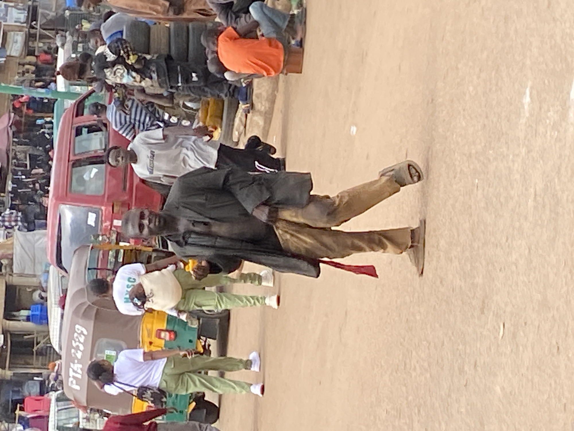 The image depicts a bustling daytime street or market scene, likely in Bukuru, Nigeria. The foreground features a man walking rightward, looking towards the viewer. He wears a dark jacket, light brown trousers, and sandals, with a strip of red fabric hanging from his hip.
Behind him, numerous people are visible, indicating a crowded environment. Two individuals wear white t-shirts paired with light green trousers; one white t-shirt prominently displays the text "MYSC" on the back. Further back, another person is seen in a bright orange t-shirt.
Vehicles include a brown motorized tricycle (keke napep) with the text "PTA 2329" visible on its rear. A red van or small bus is also parked or moving in the background amidst the crowd. The ground appears to be an unpaved, dusty surface, consistent with a street market setting.
The scene depicts everyday life with people moving about, suggesting commercial activity or general transit. The overall atmosphere is lively and active, characteristic of a busy market area.