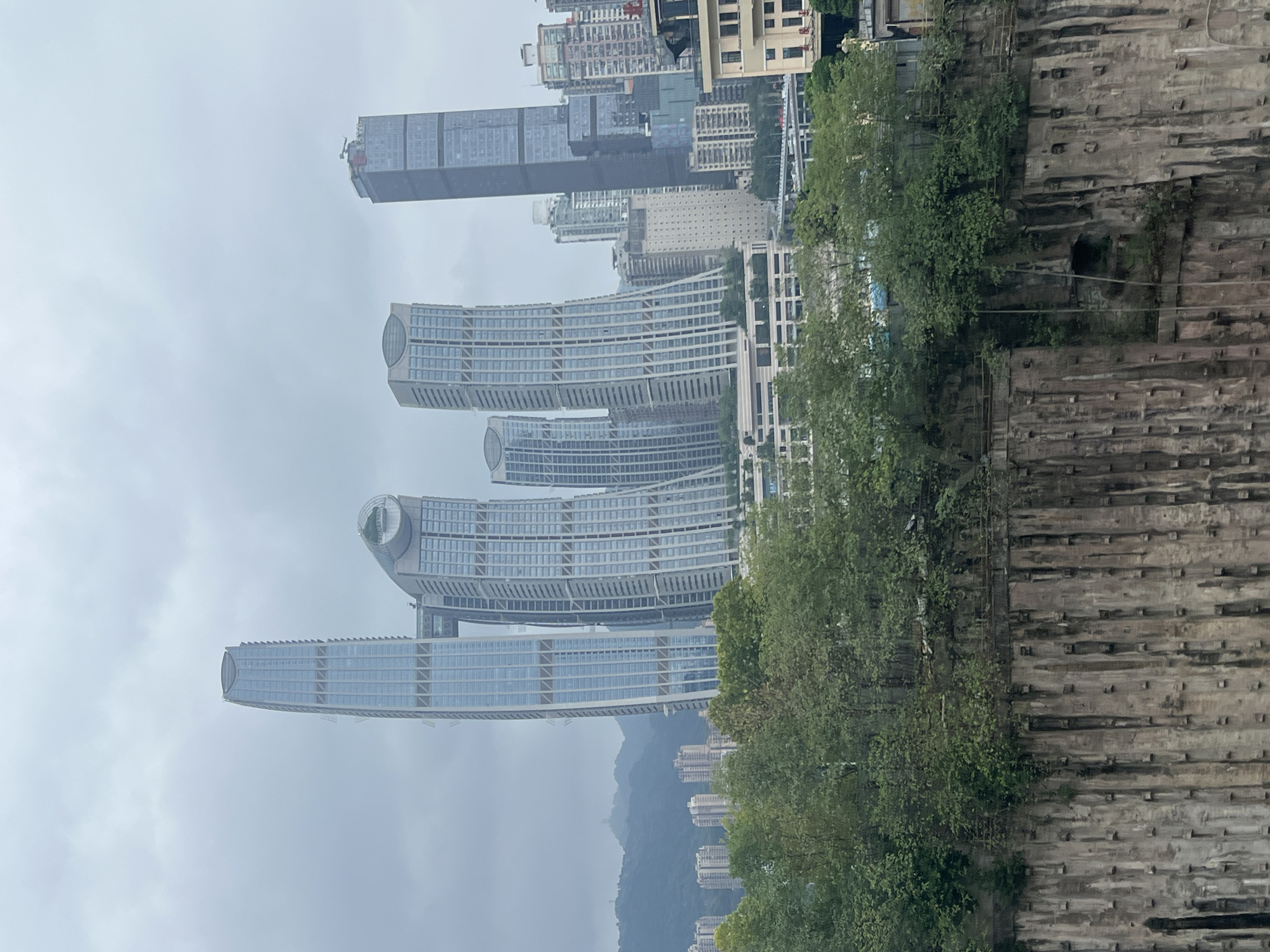 The image captures a view of the Chongqing Guotai International Building, also known as the Raffles City Chongqing, a distinctive mixed-use skyscraper complex located in Jiangbeicheng Street, Jiangbei District, Chongqing, China. The foreground is dominated by a steep, weathered cliff face with visible vertical support structures and some sparse vegetation. Above the cliff, a dense line of green trees partially obscures the lower levels of a modern cityscape.
The central focus of the image is the striking architecture of the Raffles City Chongqing. The most prominent feature is its series of interconnected, curved towers designed to resemble a sailing ship. One of these towers is particularly tall and slender, featuring a light blue glass facade with vertical white structural elements. Adjacent to it are several other similarly designed towers, some of which appear to bend or curve outwards. A horizontal skybridge, connecting two of the towers, is visible mid-way up, adding a unique architectural element.
Further in the background, a more conventional, rectangular skyscraper with a darker glass facade stands tall. Beyond these central skyscrapers, a cluster of other modern buildings of varying heights and architectural styles can be seen, indicative of a dense urban environment. In the far distance, rolling, mist-covered mountains form the backdrop to the cityscape. The sky is overcast and grey, suggesting a cloudy or possibly rainy day, contributing to a muted color palette of the scene. No people are visible in the image.