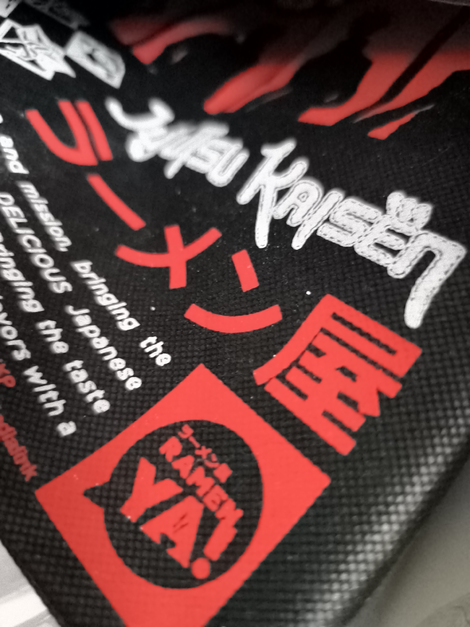 The image displays a close-up, angled shot of a black fabric material featuring red and white text and graphics. In the upper portion of the image, red flame-like shapes are visible. Below these, white lettering forms the words "Jujutsu Kaisen". To the left of "Jujutsu Kaisen", red characters are present, including a large "U" shape. Below this, white text reads "and mission, bringing the Japanese DELICIOUS the taste bringing the taste vors with a". In the lower right quadrant, a large red symbol encloses red characters that appear to be Japanese writing, with the word "RAMEN" and "YA!" in white text prominently displayed. A single person's hands are partially visible in the foreground, holding the fabric, suggesting it might be a bag or apparel. The background is out of focus and dark. There is no information available within the image to suggest a location in North Jakarta, Indonesia.