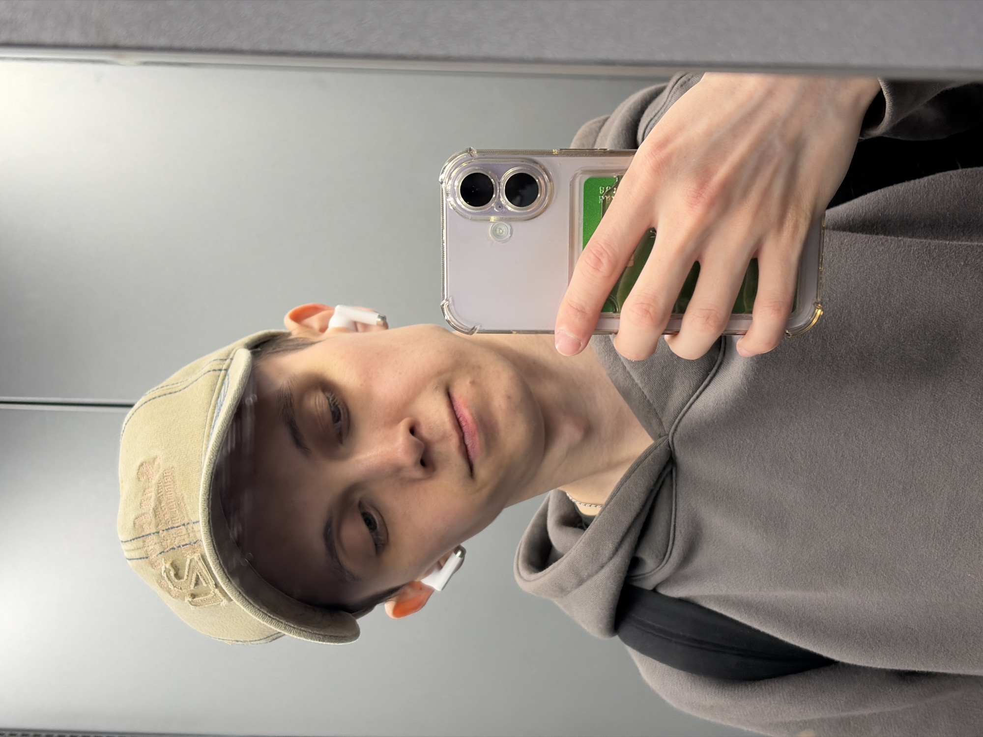 The media file is a portrait-oriented, slightly tilted selfie taken in a mirror. The main subject is a person with short, light-colored hair, wearing a light tan baseball cap and a grey hooded sweatshirt. They have white earbuds in both ears. The person is holding a smartphone in their right hand, which is capturing the reflection. The phone has a clear protective case with a green card visible inside.
The background is a plain, neutral grey wall, suggesting an indoor setting. The lighting appears to be soft and diffused, with no strong shadows, indicating it could be daytime or well-lit indoor conditions. The expression on the person's face is neutral or slightly amused.
The image displays no discernible text, apart from what might be on the phone's screen, which is not clearly legible, or on the baseball cap, which is also difficult to read clearly. The overall impression is a casual, everyday moment captured by the subject.