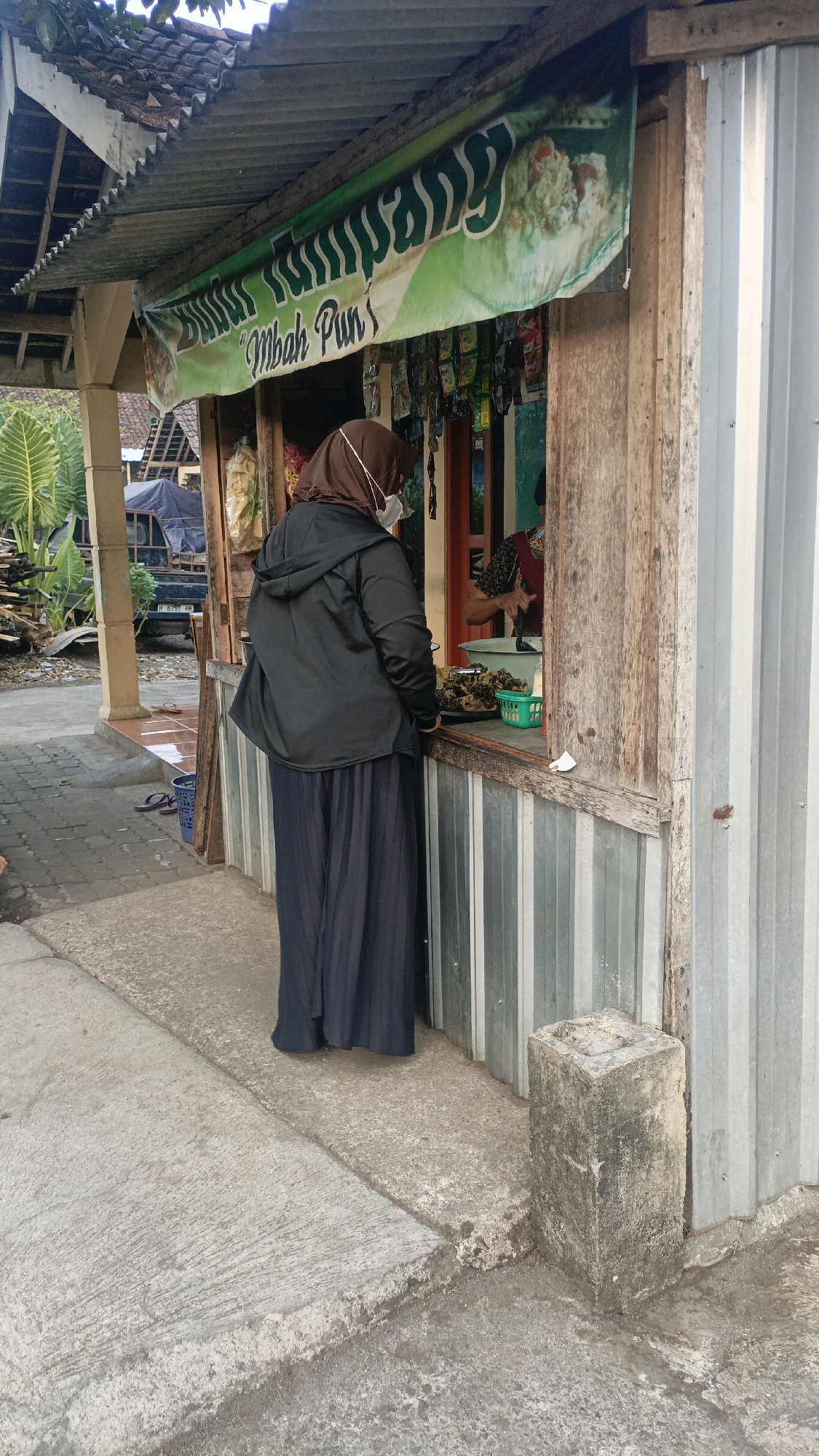 A woman wearing a hijab and face mask stands at a food stall, facing a window. She is dressed in a black hooded jacket and a dark blue pleated skirt. The stall has a corrugated metal facade and a wooden counter. Above the counter, a green banner with Indonesian text and images of food is displayed. Various food items are visible on the counter, along with a green basket. Another person, partially visible behind the counter, appears to be preparing food. The stall is situated on a concrete ground. In the background, a truck and some vegetation are visible. The scene is set in Salatiga, Indonesia, indicated by the style of the stall and the language on the banner.