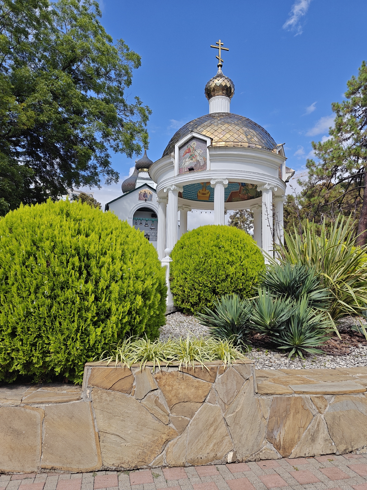 This is a photograph of a white gazebo with a golden dome topped with a cross, situated in front of a church with a similar dome. The gazebo has ornate columns and mosaic art on its exterior. In the foreground, there are meticulously landscaped bushes and spiky plants, with a stone wall bordering the area. The scene is set against a clear blue sky with scattered clouds, and lush green trees frame the background. The location appears to be an outdoor religious site, possibly a churchyard or garden, with a focus on architectural beauty and serene greenery.