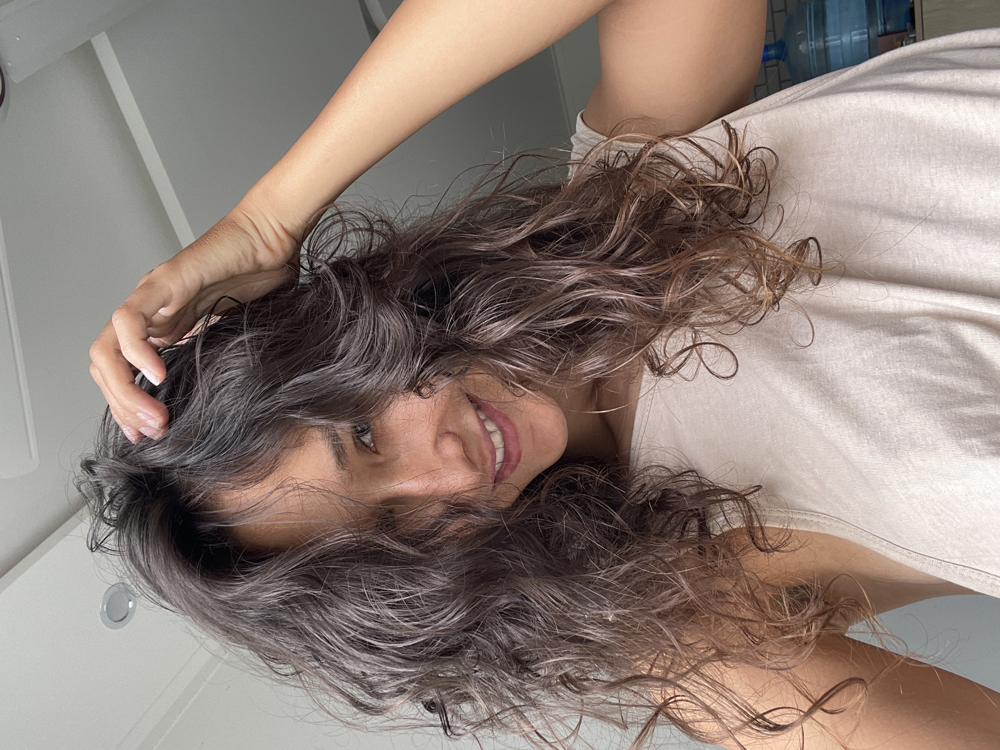 A woman with long, wavy brown hair is smiling at the camera. She is wearing a light-colored t-shirt and has one hand touching her hair. The background is a white wall and ceiling, with a glimpse of what appears to be water bottles in the upper right corner. It is impossible to confirm the location as Mui Ne, Vietnam from this image alone.
