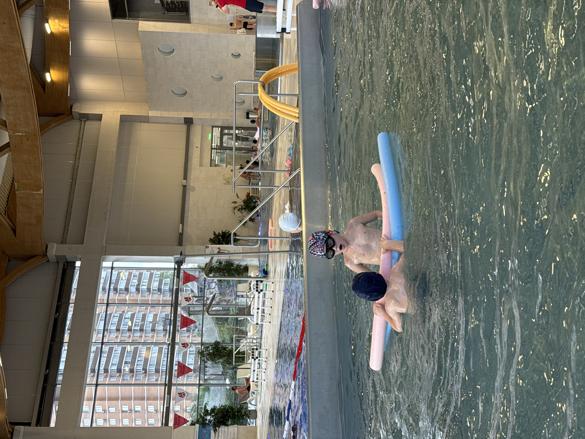 Two young boys are in a swimming pool, one wearing a black swim cap and the other wearing a patterned swim cap and goggles. They are holding onto a pink pool noodle. The pool is indoors with a high ceiling and large windows that look out onto buildings and trees. The environment appears to be a public swimming facility, with other people visible in the background, some relaxing on lounge chairs. The water is clear and shows ripples from activity.