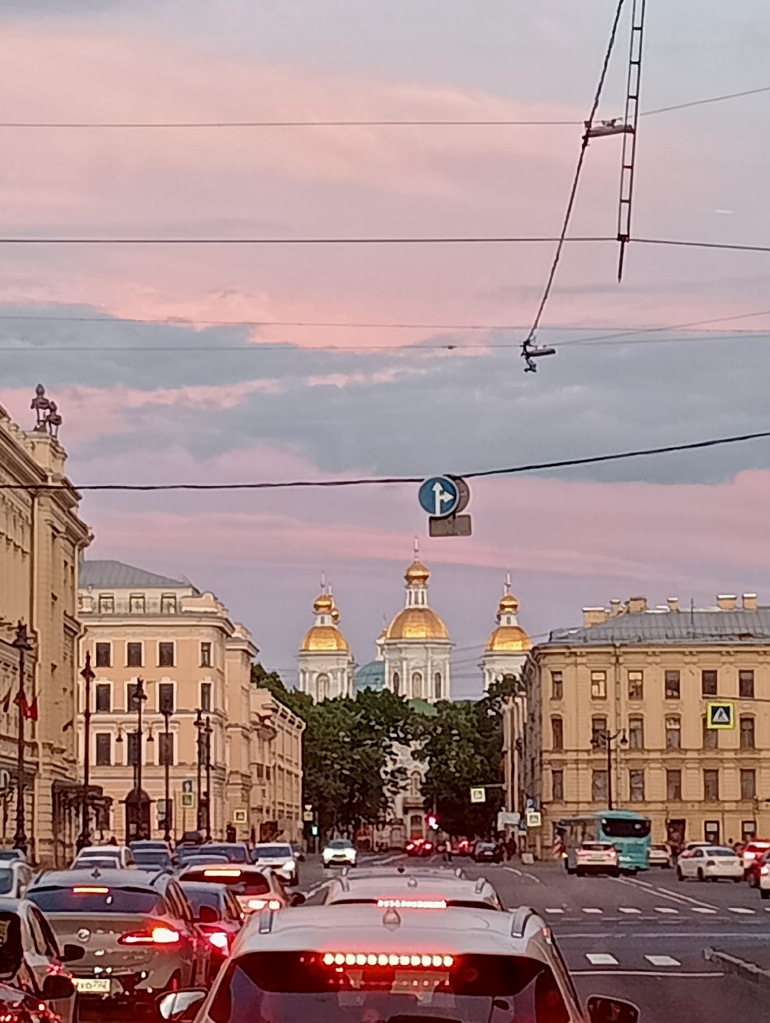 The image captures a traffic jam on a street in Saint Petersburg, Russia, with a distinctive pink and purple sky overhead. In the background, the golden domes of Saint Nicholas Naval Cathedral are visible, a prominent landmark in the city. The architecture of the surrounding buildings is typical of Saint Petersburg, with classical facades and ornate details. Cars are stalled on the road, their brake lights illuminated. Overhead, power lines crisscross the sky, and a traffic sign indicating a turn is visible. The overall scene depicts a bustling urban environment with a serene, atmospheric sky.