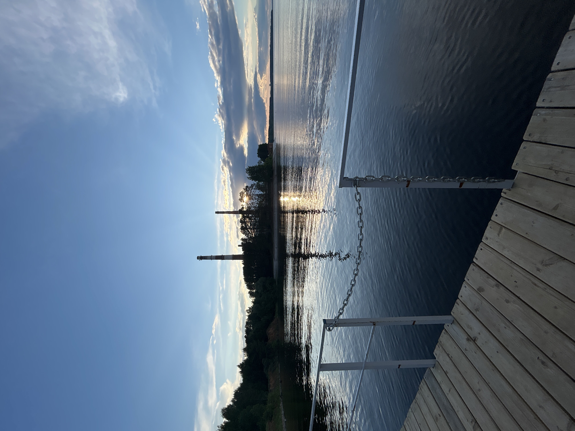 The image shows a scene in the city of Chaikovsky, Russia. From a wooden dock, the view looks out over a body of water, likely a lake or river. Two tall industrial smokestacks are visible on the opposite bank, silhouetted against the setting sun, which is reflected in the water. The sky is blue with scattered clouds. Metal railings and a chain are attached to the dock. Trees line the bank across the water. The sun's rays are visible through the clouds.