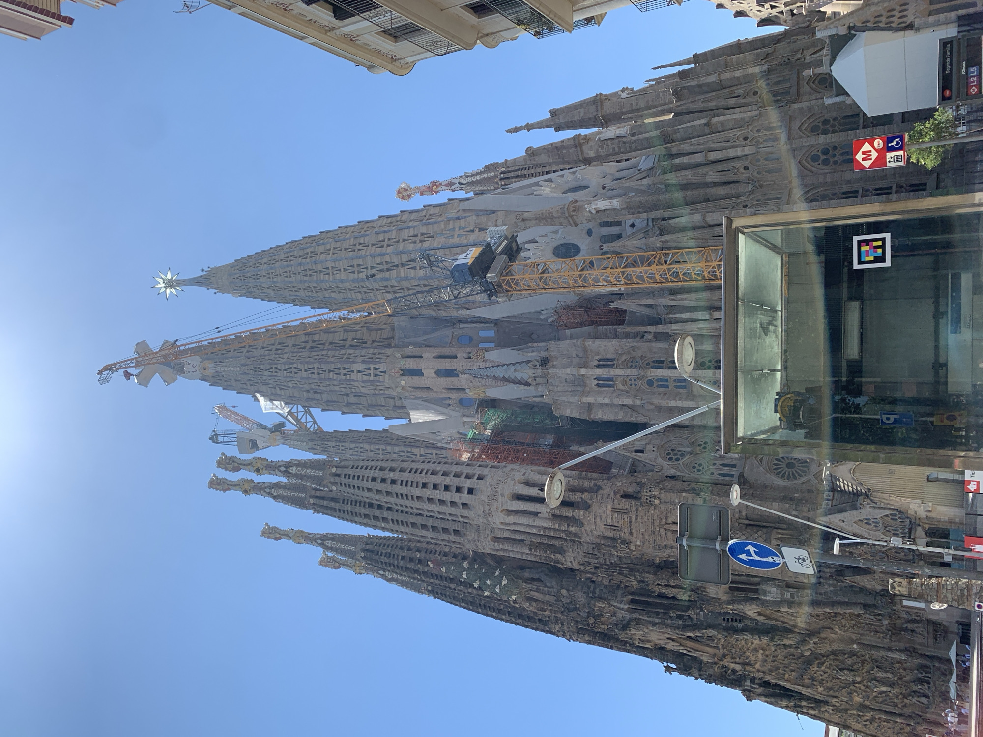 The image displays a low-angle, full shot of the Sagrada Familia basilica in Barcelona, Spain. The iconic, unfinished cathedral dominates the frame with its intricate facade and towering spires. Several construction cranes are visible, indicating ongoing work on the basilica. The sky is clear and blue, with the sun casting a bright light, creating some lens flare. In the foreground, there is a modern glass structure, likely an entrance or information booth, with signage. A street light and traffic signs are also present. The surrounding buildings, visible on the right and left edges of the frame, are typical of Barcelona's urban architecture. No people are clearly discernible in the foreground or middle ground, though there may be some individuals present in the distant background on the sidewalk. The overall scene depicts a sunny day at a major tourist attraction undergoing construction.