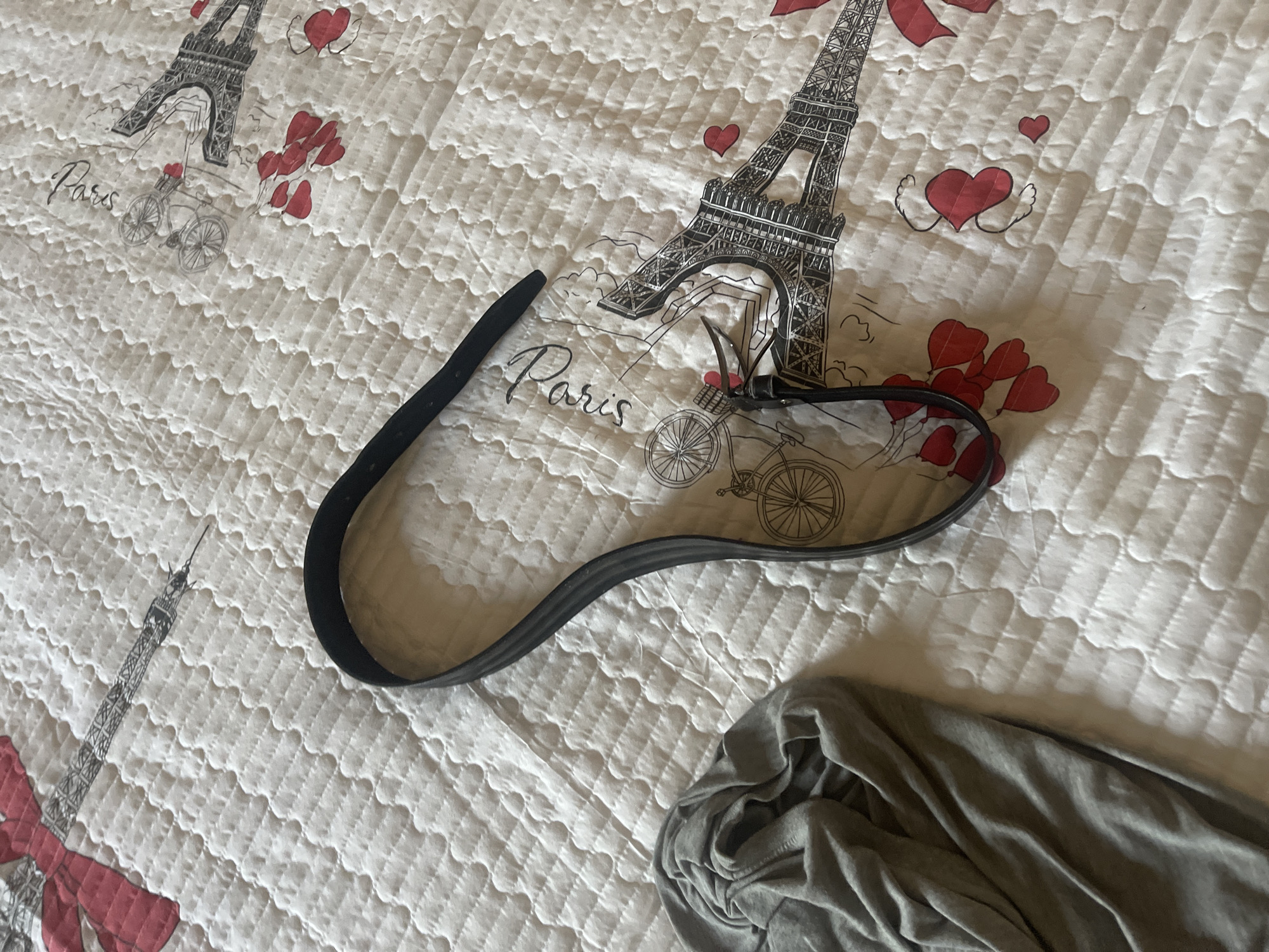 A black belt lies in the center of the frame, looped into a horseshoe shape. To the left of the belt, a crumpled grey garment is visible. The background is a white quilted blanket featuring a pattern of Parisian landmarks, including the Eiffel Tower, hearts, and the word "Paris" written in script. There is no discernible connection to the city of Qarshi, Uzbekistan, as the image depicts domestic items on a blanket.