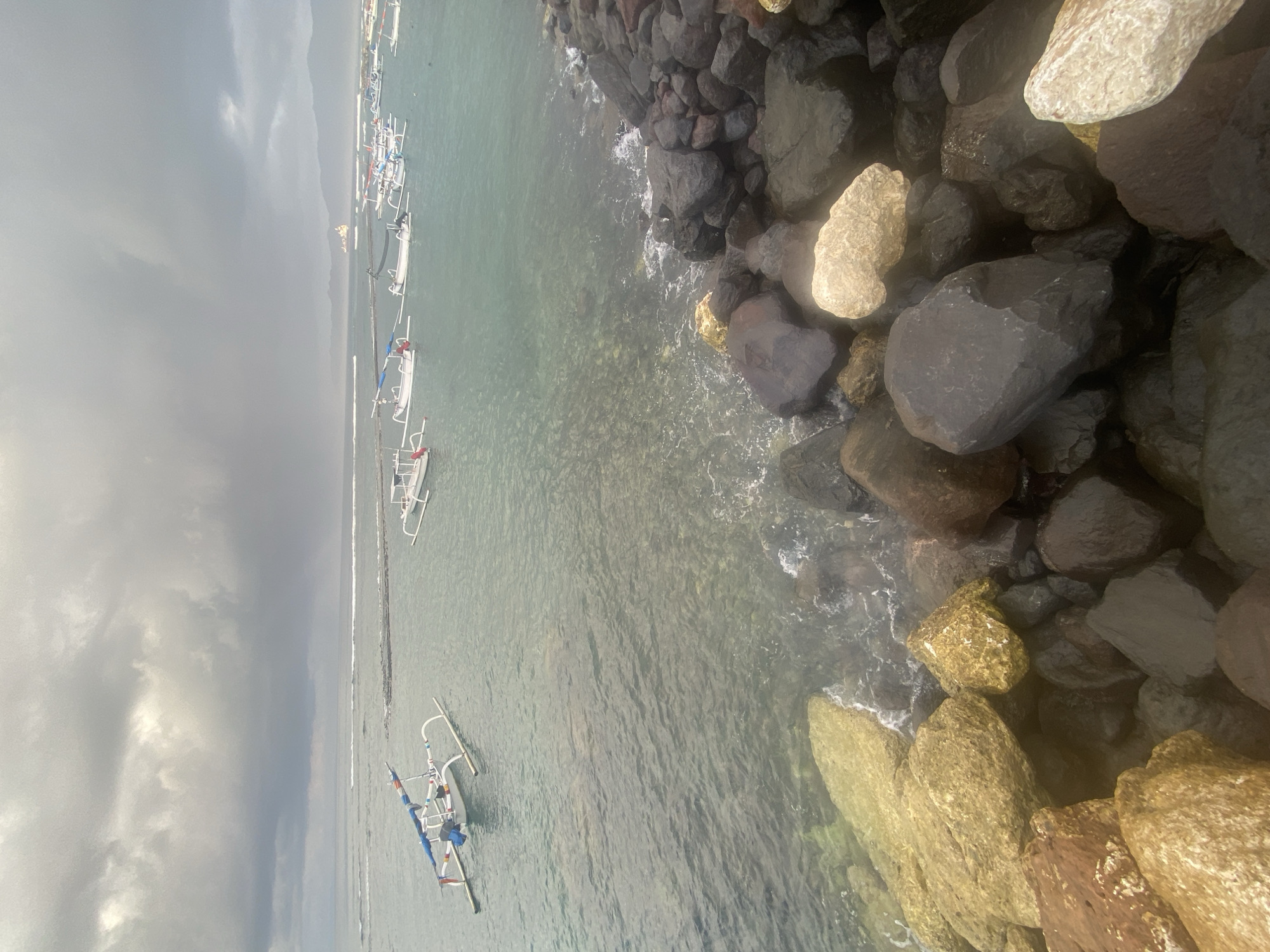 This image shows a coastline scene in Karangasem, Indonesia. The foreground features a rocky shore with large, dark and light-colored boulders. The water is a clear, greenish-blue, with small waves lapping against the rocks. In the middle ground, several traditional Indonesian outrigger boats are moored. These boats are white with colorful accents, and some have blue canopies. The water in this area appears shallow enough to see the seabed in places. In the background, the sea stretches out towards a hazy horizon, with a few more boats visible further out, and a larger vessel, possibly a cargo ship, in the distance. The sky is overcast with dramatic, dark clouds, suggesting a moody or potentially stormy weather condition. The overall impression is a tranquil, yet somewhat somber, coastal landscape.
