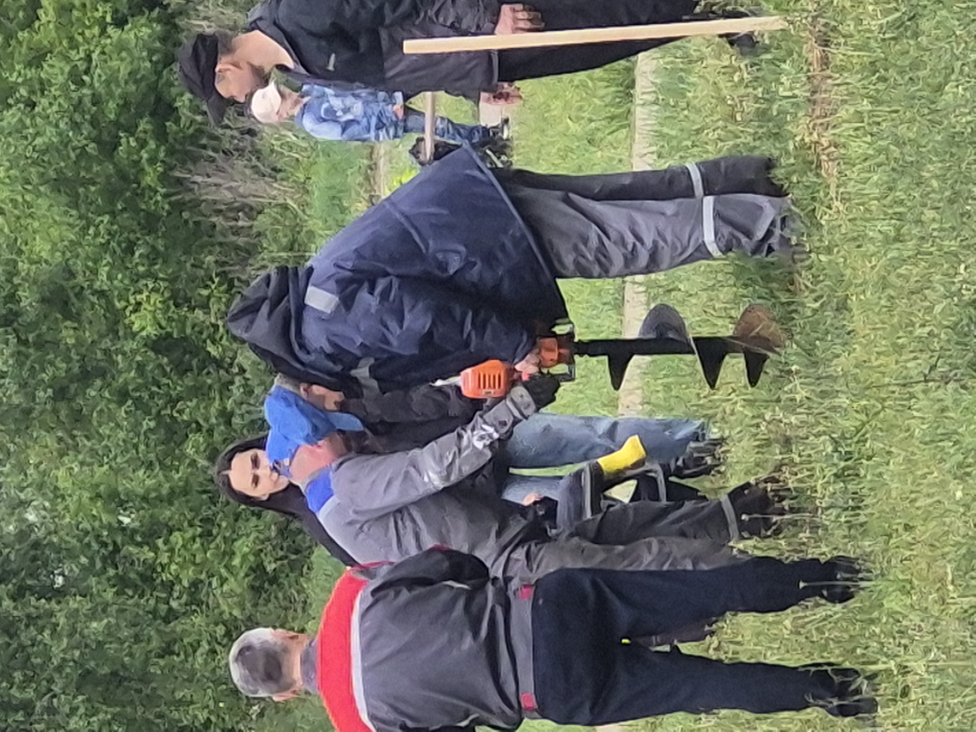A group of people are standing on a grassy field. One person is operating an orange auger. Some people wear black clothes. One person holds a wooden stick. Another wears a red and grey jacket. The city is Unknown, Unknown.