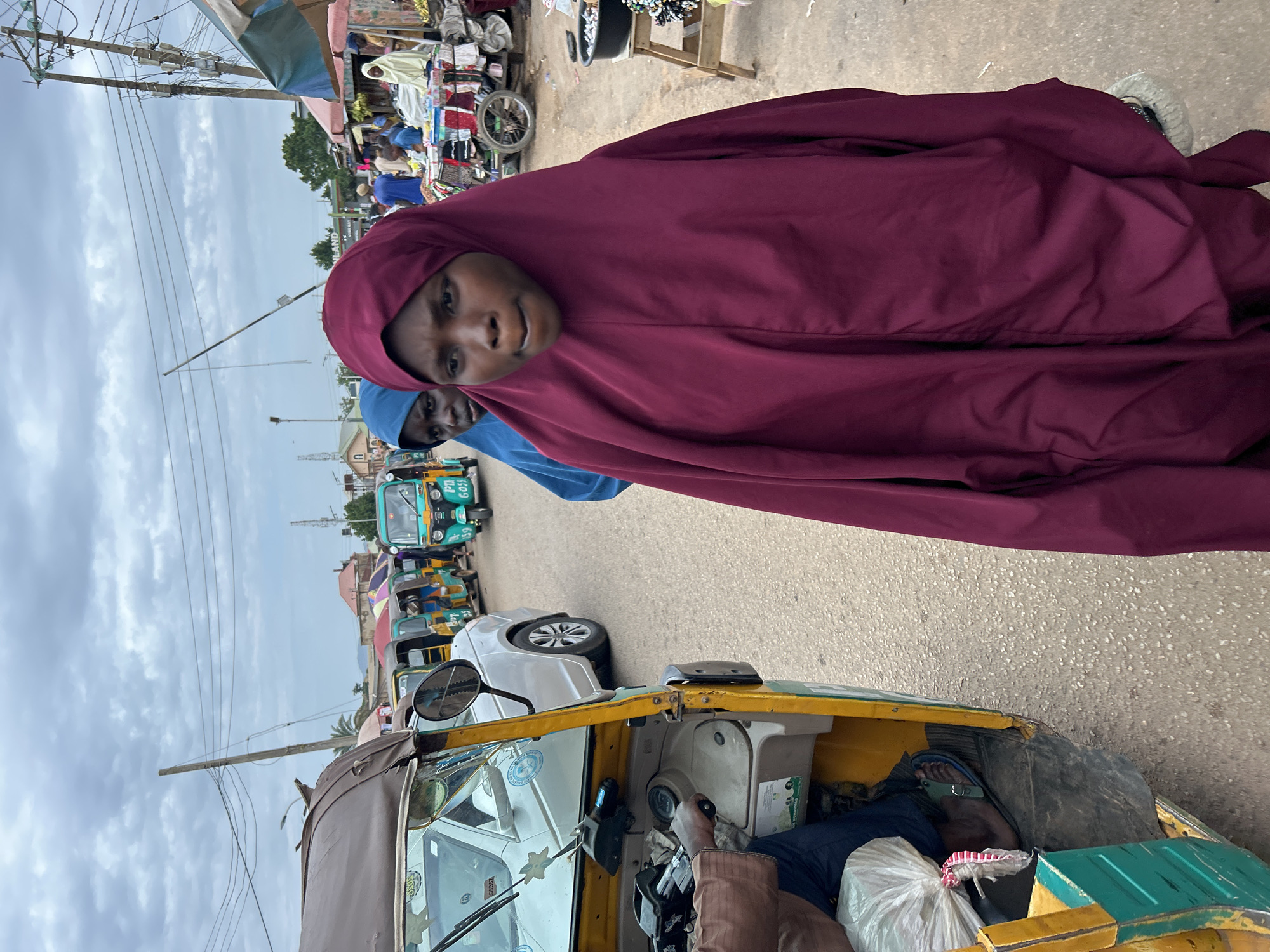 The image captures a street scene in Jos, Nigeria, likely in a market area. In the foreground, two young women are walking towards the camera, both wearing hijabs. The woman on the right is closer to the viewer and is wearing a deep red hijab that covers her entire body. Her face is partially obscured, but her mouth is open as if she is speaking. The woman on the left is wearing a bright blue hijab and is looking slightly away from the camera.
To the left of the women, a yellow and green auto-rickshaw, commonly known as a 'Keke Napep', is parked. The driver's arm and hand are visible on the steering wheel. In the background, a line of similar auto-rickshaws are parked or moving along the street. There are also market stalls visible with various goods displayed under colorful awnings. The sky is overcast, suggesting a cloudy day. The overall impression is one of a bustling urban environment in Nigeria.