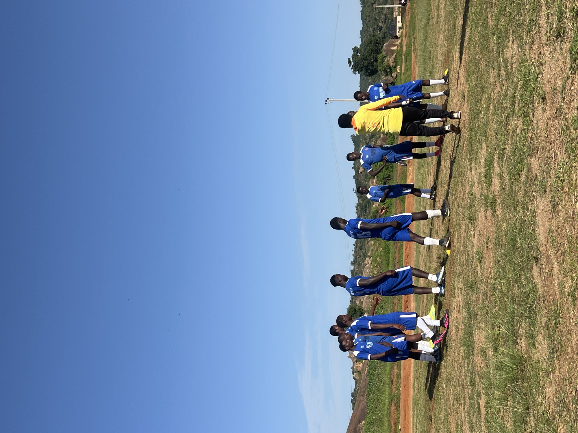 A group of young Black men in soccer uniforms are standing on a grassy field under a clear blue sky. They are lined up in two rows, facing the same direction. Most are wearing blue jerseys and shorts with white socks, but one person in the back is wearing a yellow jersey and black pants. The field appears dry and somewhat patchy, with some exposed dirt. In the background, there are hills with sparse vegetation and a few buildings. A power line pole with insulators is visible in the mid-ground. The lighting suggests it is daytime, likely late morning or early afternoon, given the bright sunlight and distinct shadows. The individuals appear to be attentive and possibly engaged in a training session or a pre-game huddle. No overt emotions are discernible from their stances or facial expressions. The scene is set in Jos, Nigeria, as indicated by the context.