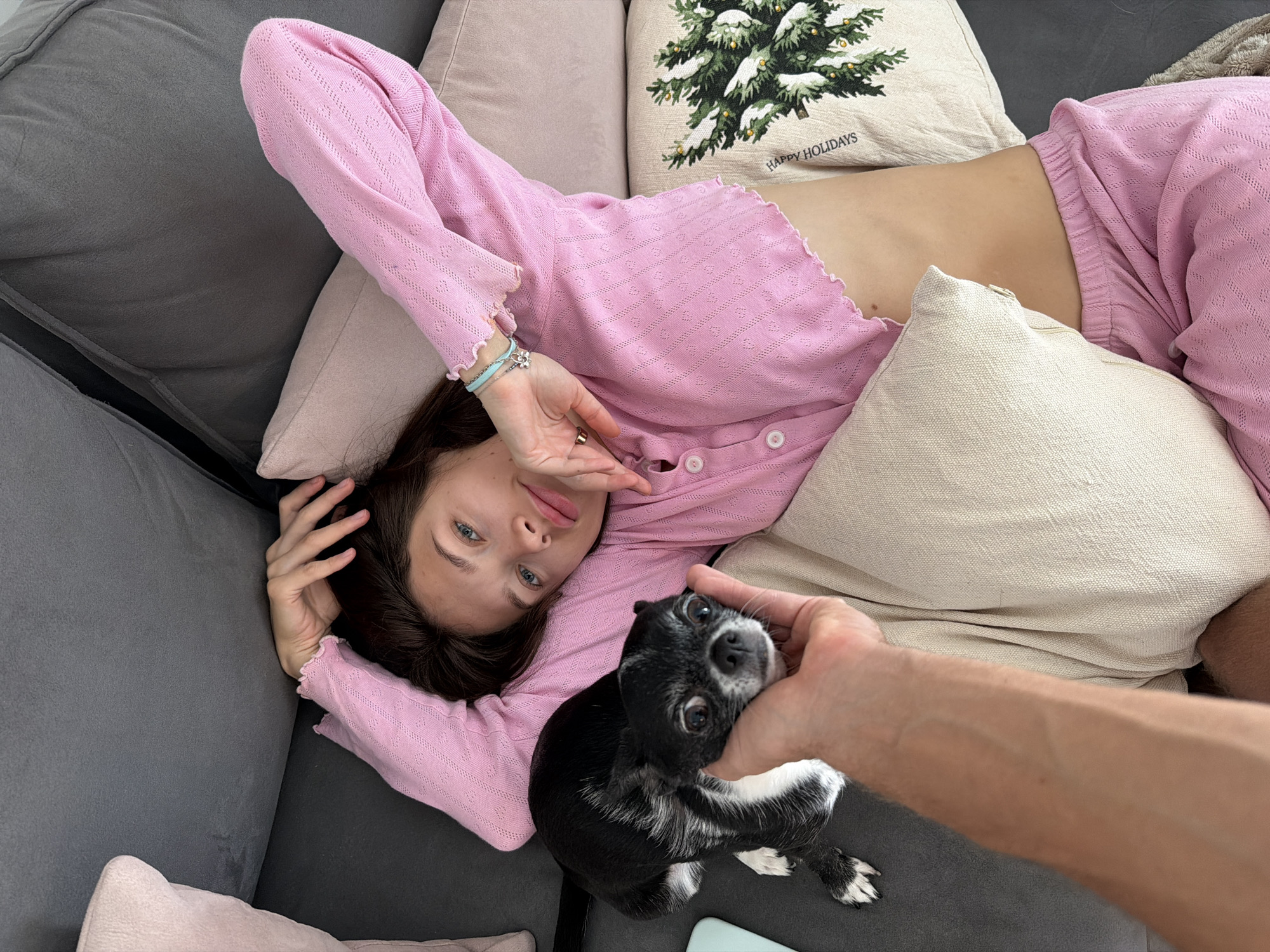 A woman in a pink outfit reclines on a gray couch, surrounded by several pillows. One pillow features a green Christmas tree design with the words "HAPPY HOLIDAYS." A small black and white dog is held by a person's hand, positioned near the woman. The location appears to be indoors, specifically on the couch.