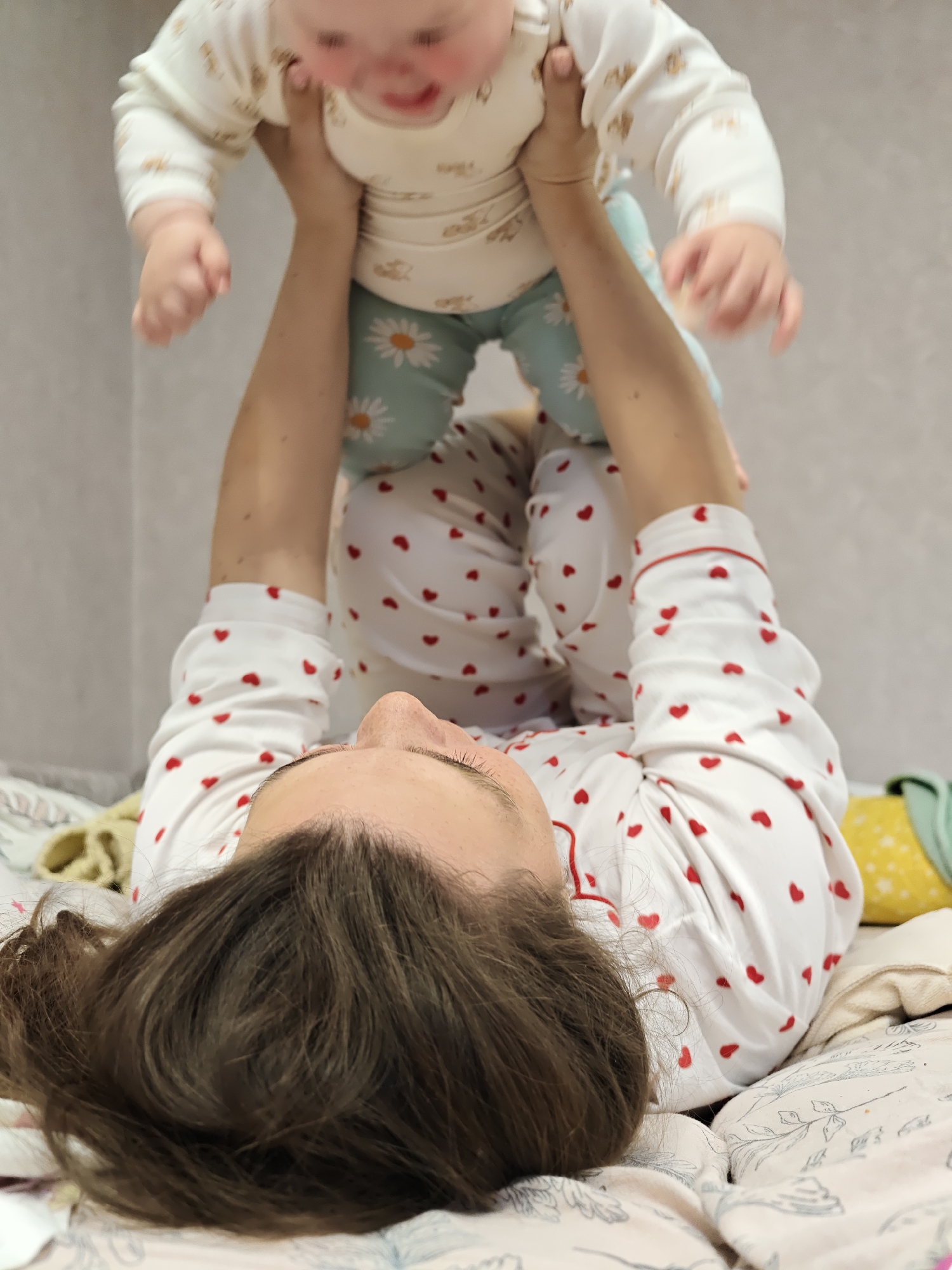 A woman lies on her back on a bed covered with a patterned sheet, wearing a white pajama top with red hearts. She holds a baby up in the air above her, the baby wearing a patterned onesie and pants with daisy prints. The location is suggested to be Chaikovsky, Russia, but no visual details in the image confirm this.